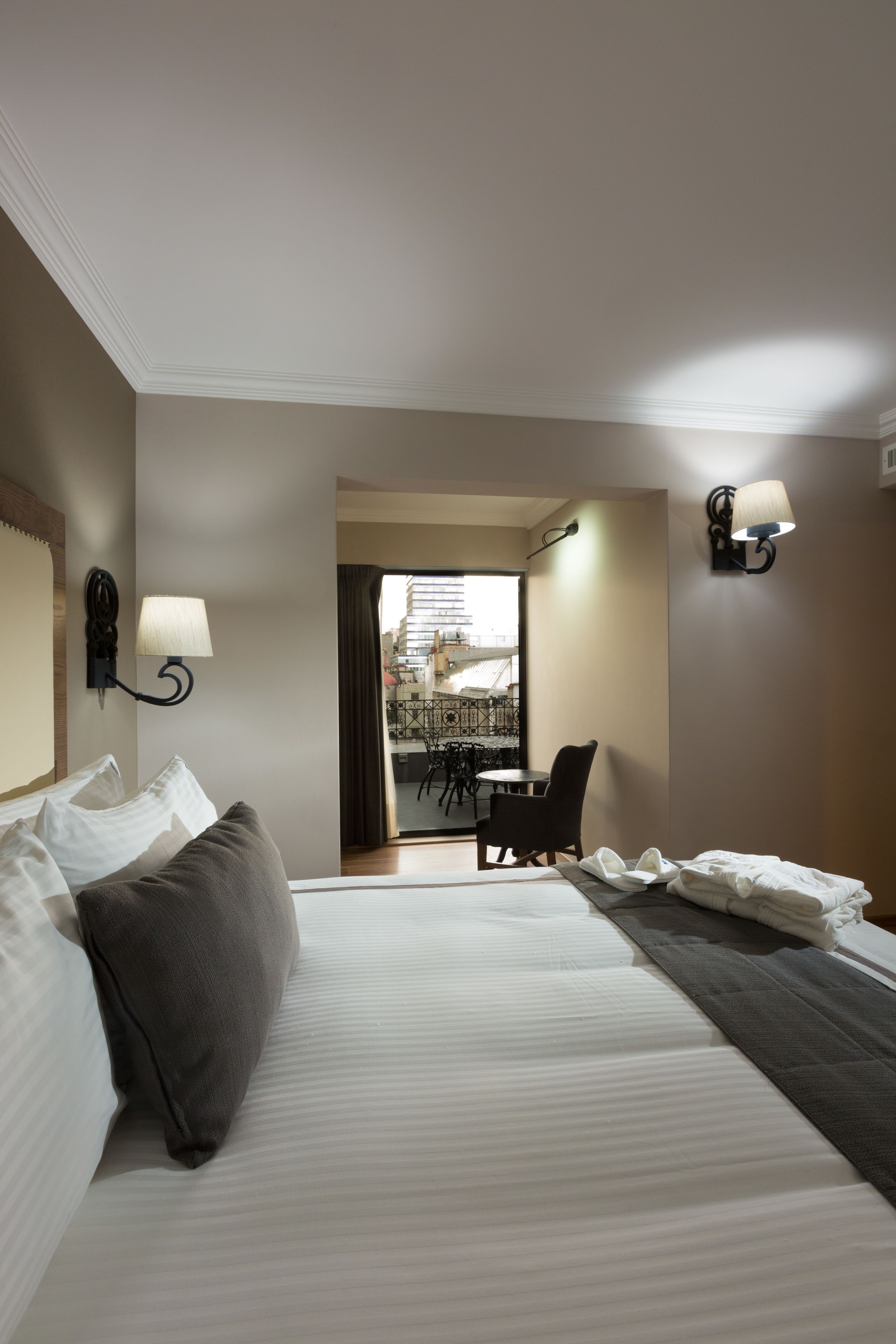 A hotel room with a bed, two wall-mounted lamps, a view of the city through a door, and a balcony with a table and chairs.