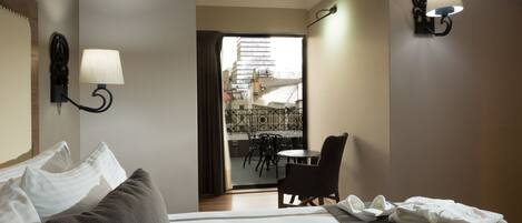A hotel room with a bed, two wall-mounted lamps, a view of the city through a door, and a balcony with a table and chairs.