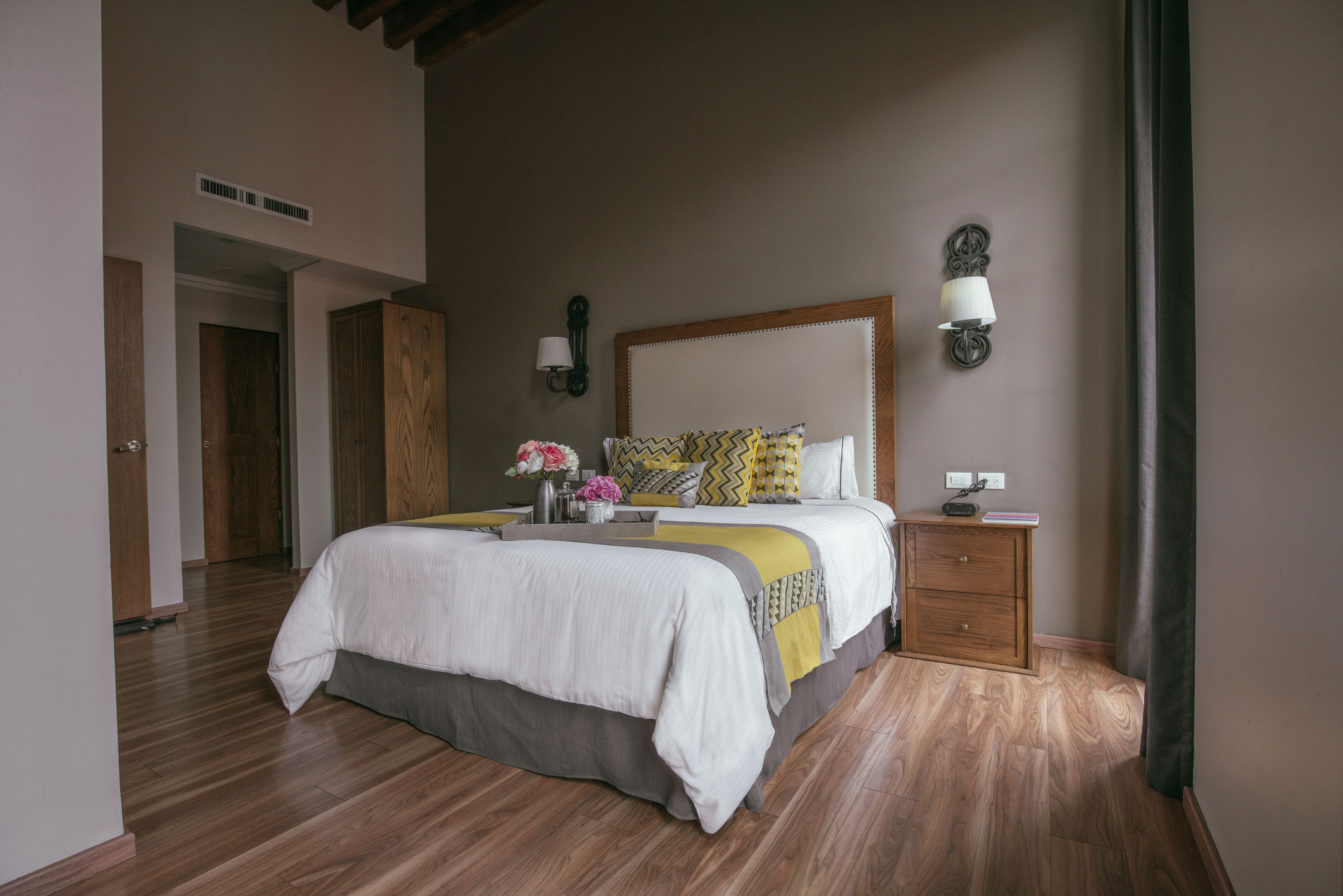 Quarto com cama grande, piso de madeira, mesa de cabeceira e luminária na parede.