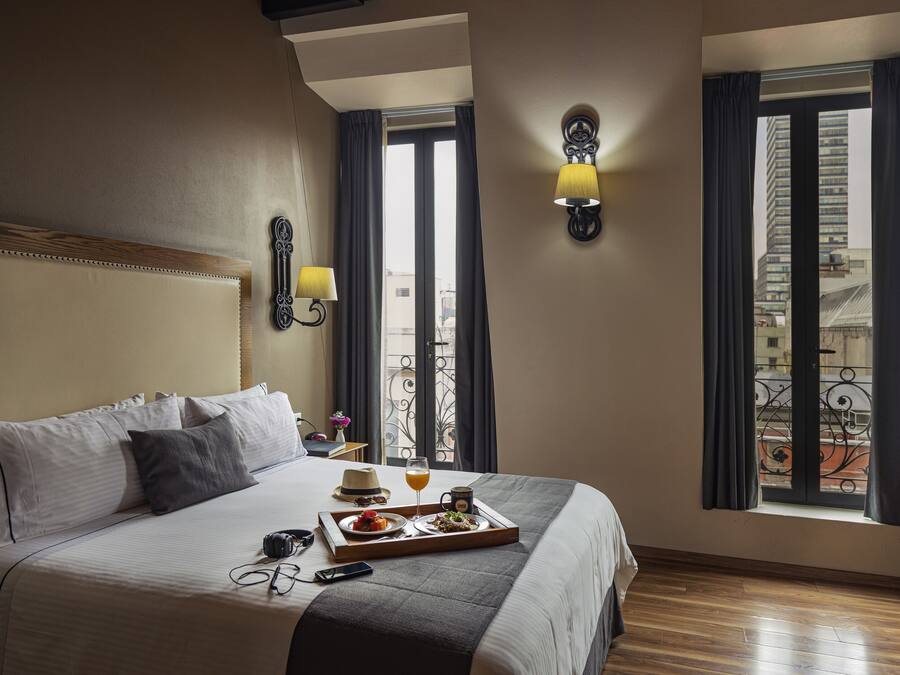 A hotel room with a large bed, a tray with breakfast, a laptop, and a view of the city through the window.