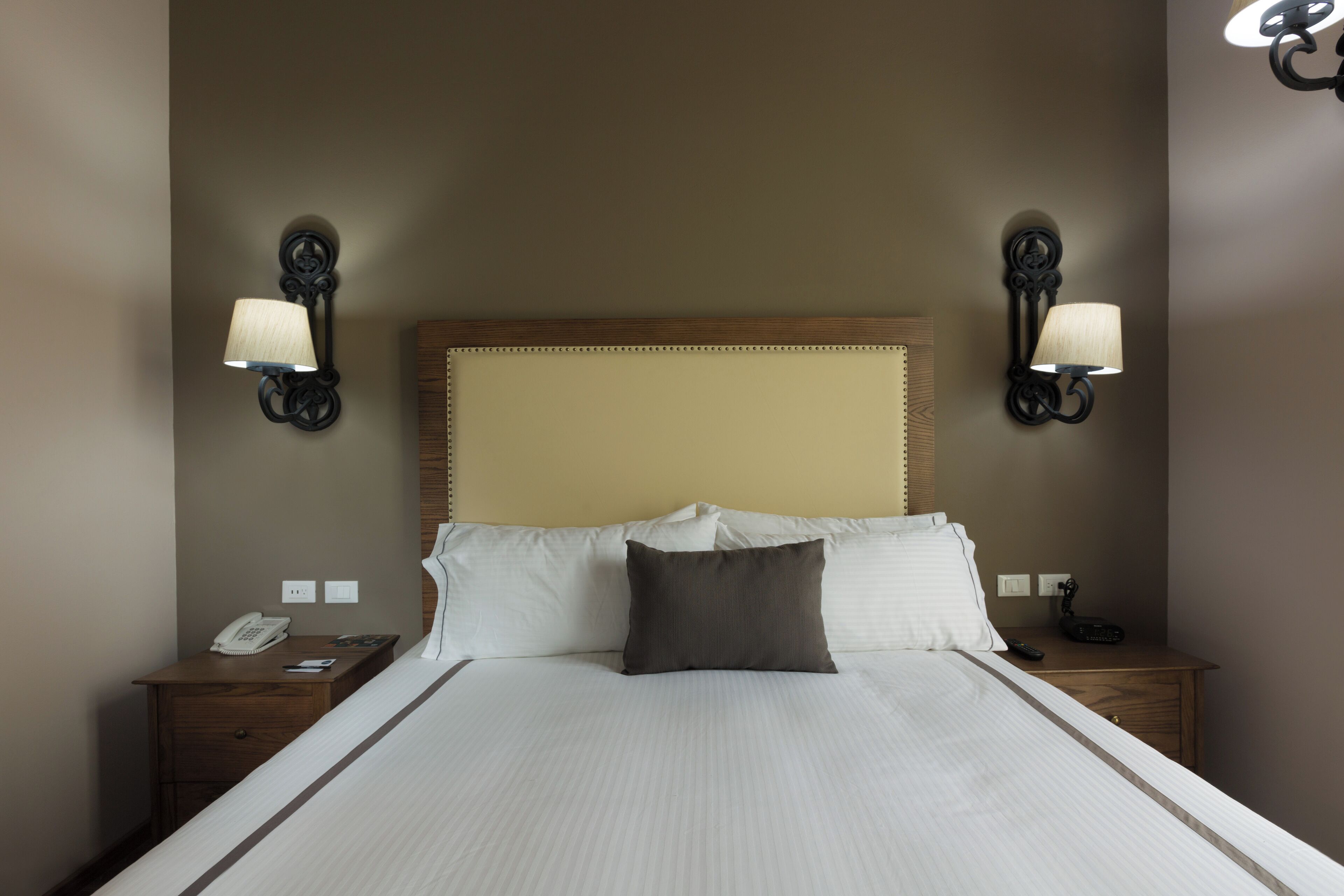 Una cama bien hecha con cabecera de madera, dos mesitas de noche con lámparas y un teléfono sobre la de la izquierda.