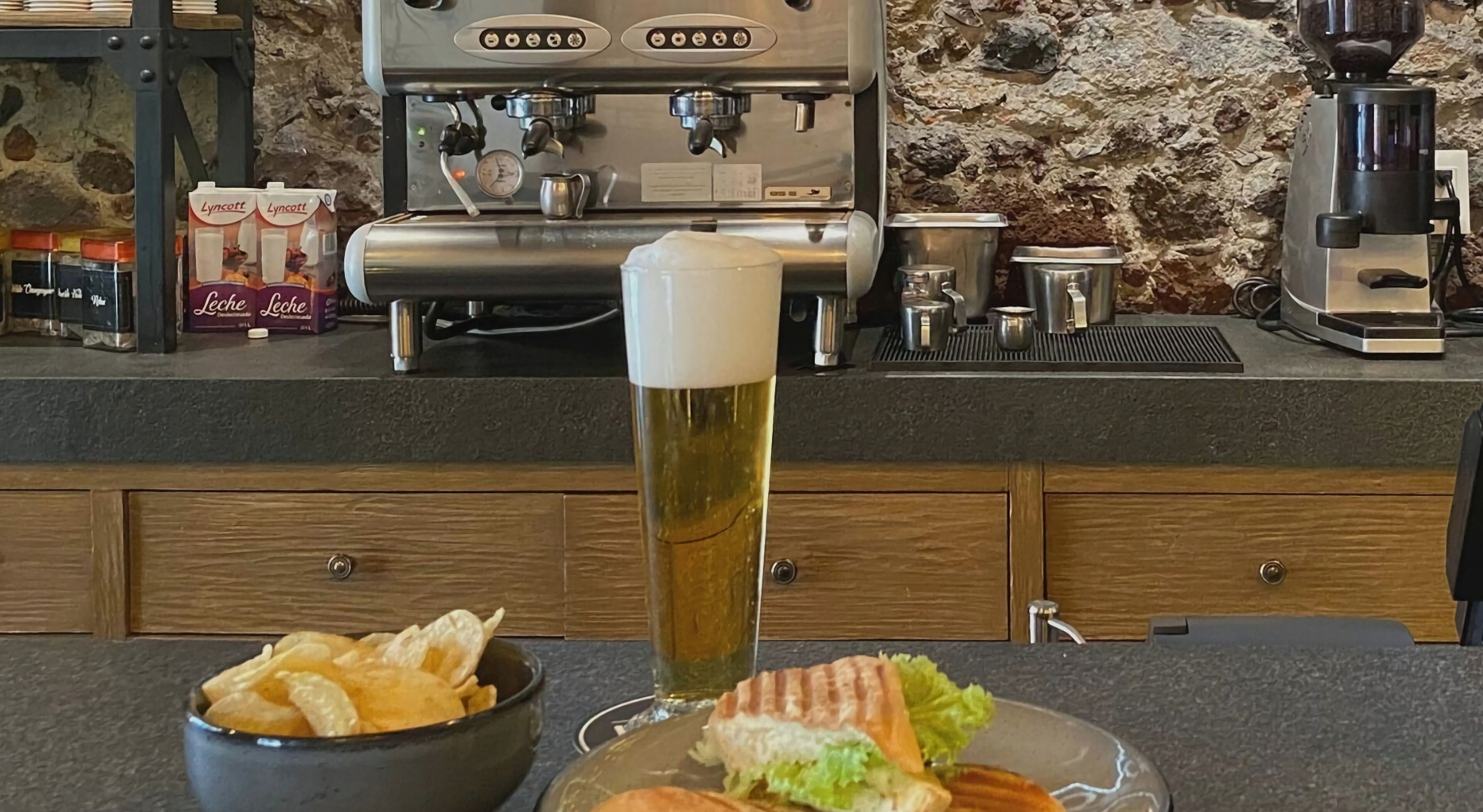 A bar area with a tall beer glass, a bowl of chips, and a sandwich on a table.