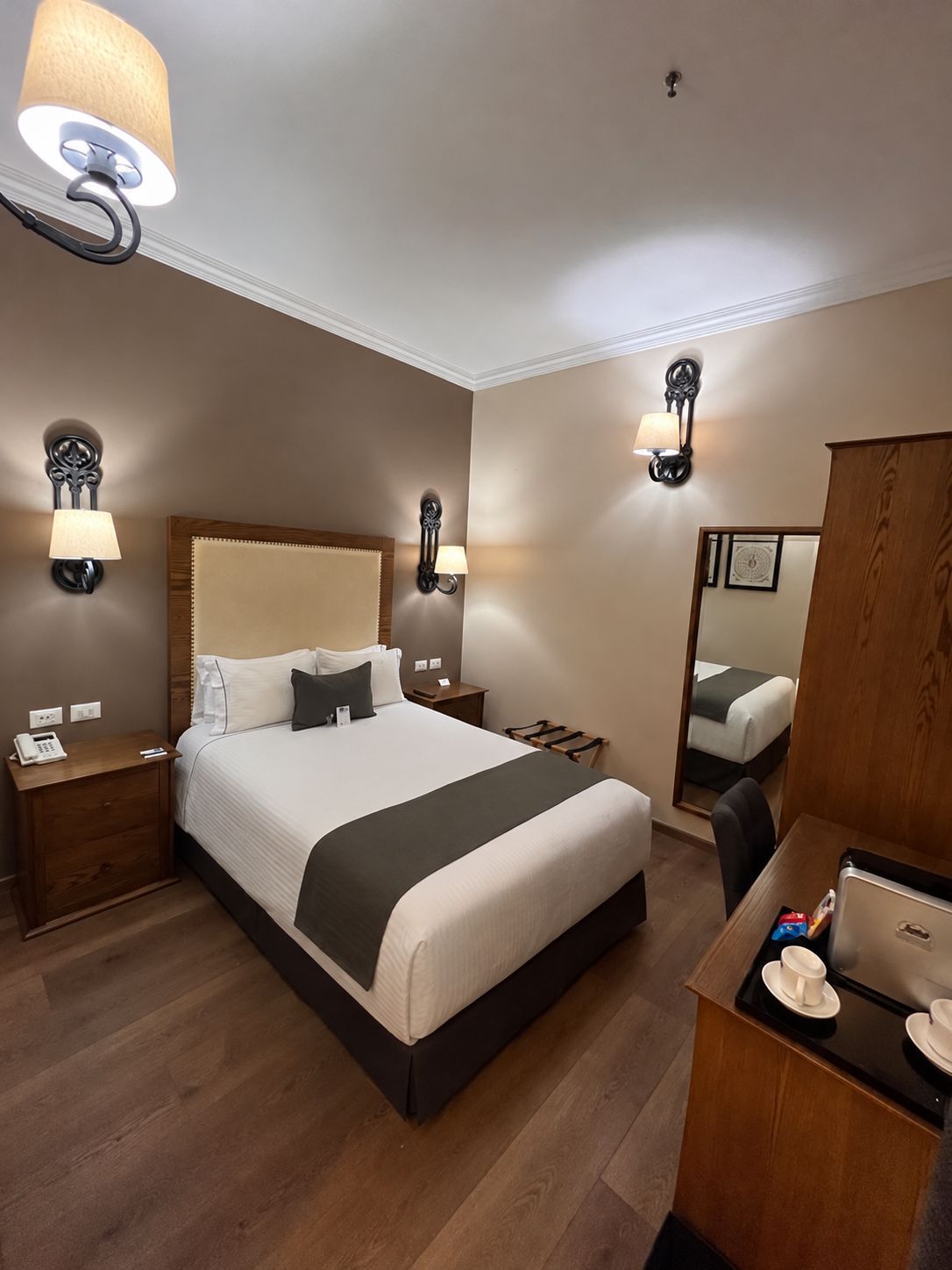 A hotel room with a large bed, bedside tables, a desk with a computer, and a mirror.