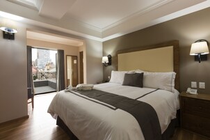 A hotel room with a large bed, a wooden nightstand, a lamp, and a view of the city through a glass door.