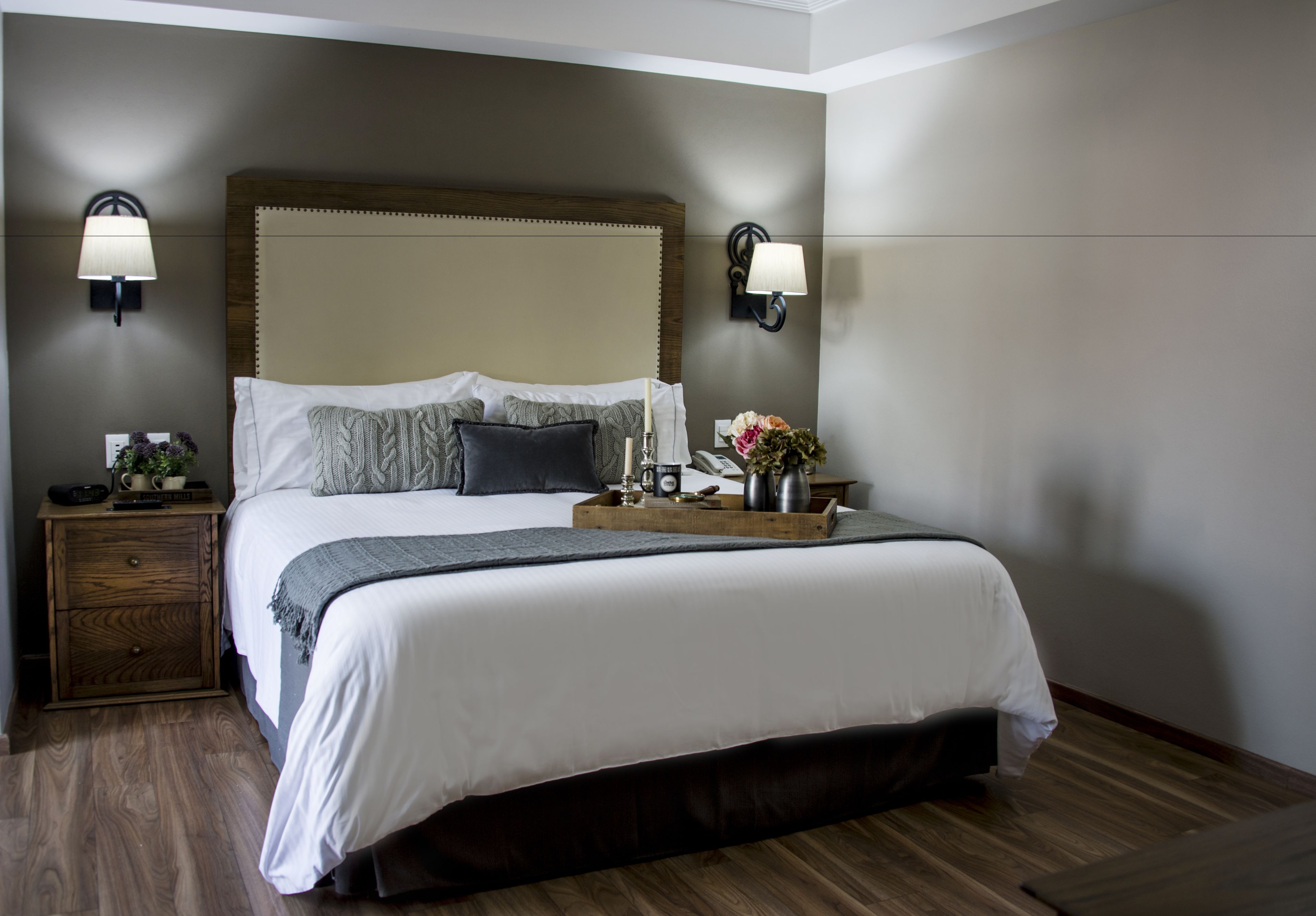 Una cama bien hecha con cabecera de madera, dos lámparas de noche, una mesita de noche con lámpara y una bandeja con flores y un florero.