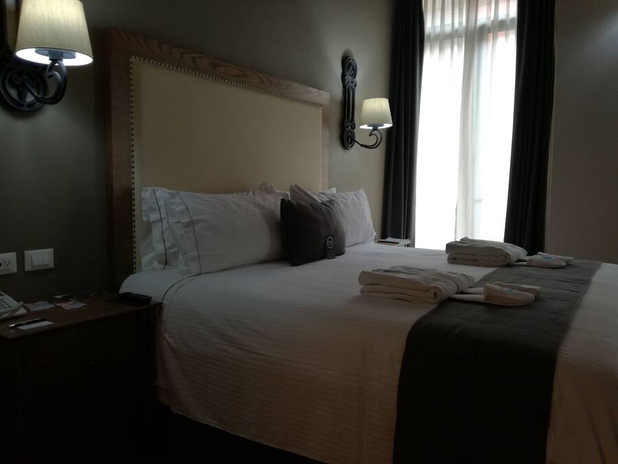 Habitación de hotel con una cama grande, dos lámparas de noche, una mesita de noche con un teléfono y una ventana con cortinas.