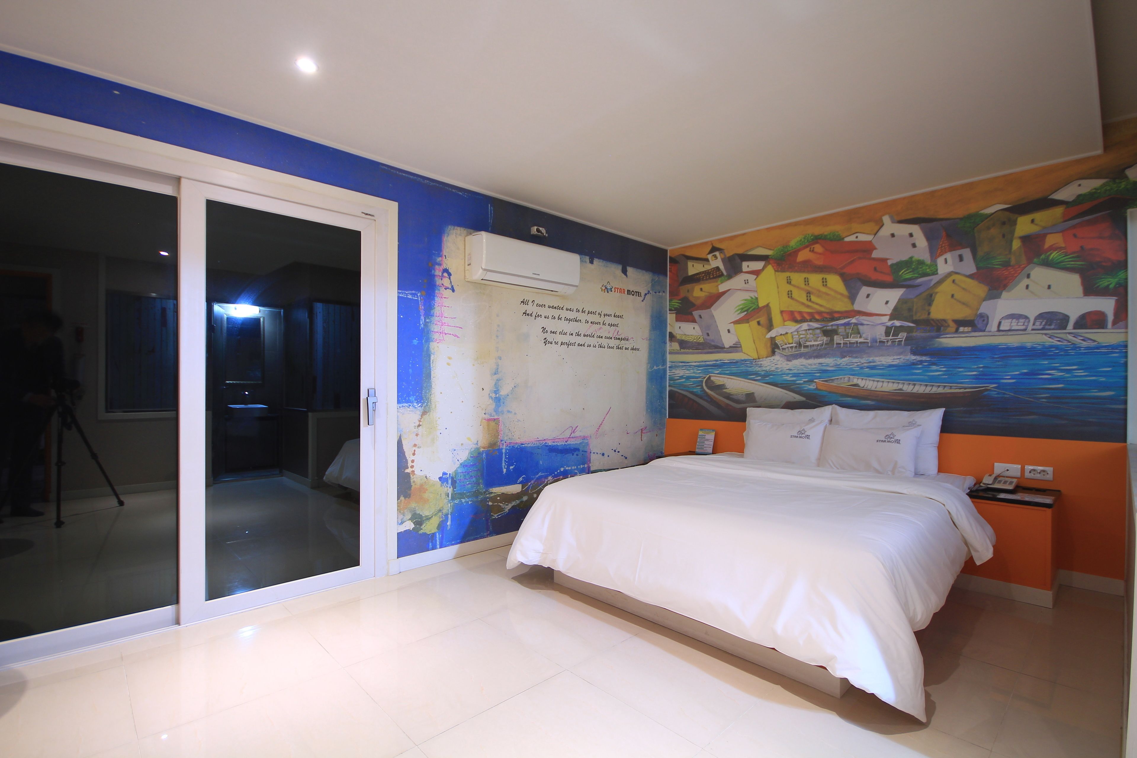 Quarto com uma cama grande, porta de vidro deslizante para uma varanda, um mural colorido na parede e uma mesinha com um telefone e um abajur.