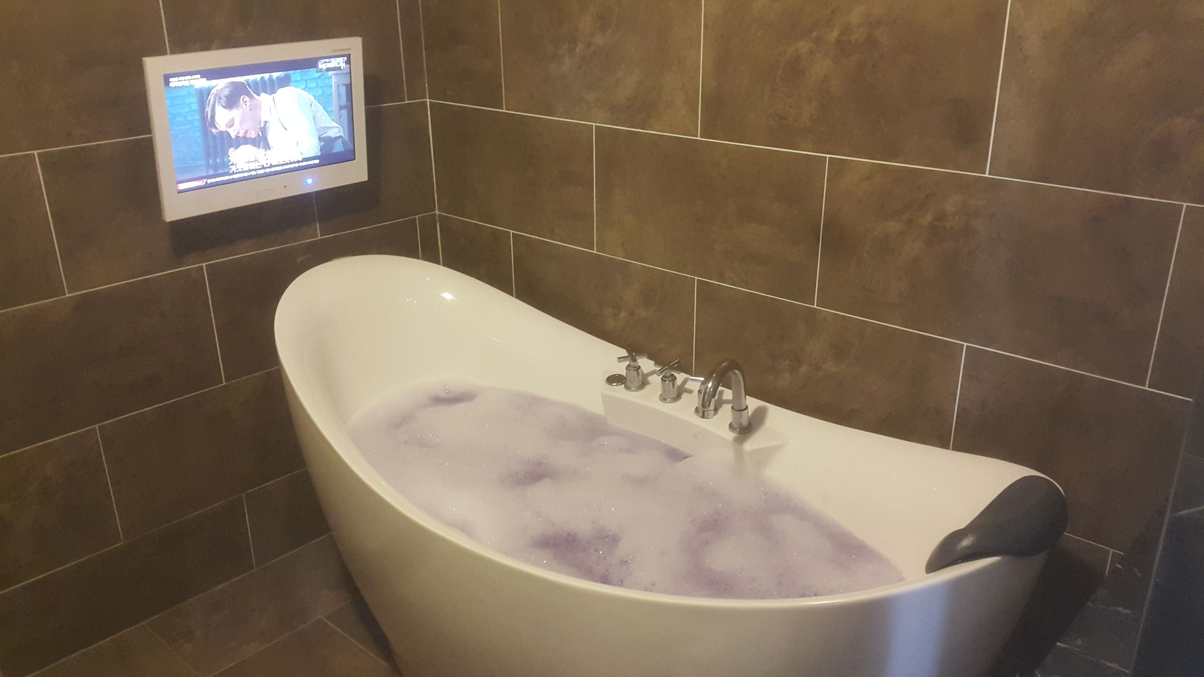 A bathtub with bubbles, a TV mounted on the wall, and brown tiled walls.