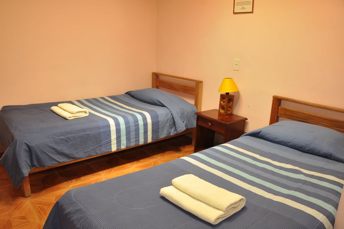 Two single beds with blue striped bedding and white towels, a wooden nightstand with a yellow lamp, and a wooden floor.