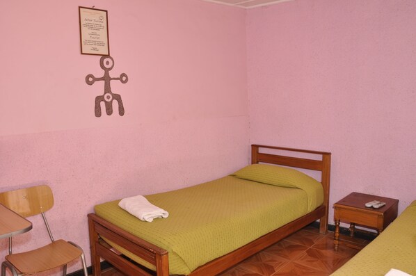 Una habitación pequeña con paredes rosadas, una cama individual, una silla de madera, una mesita y un certificado enmarcado en la pared.