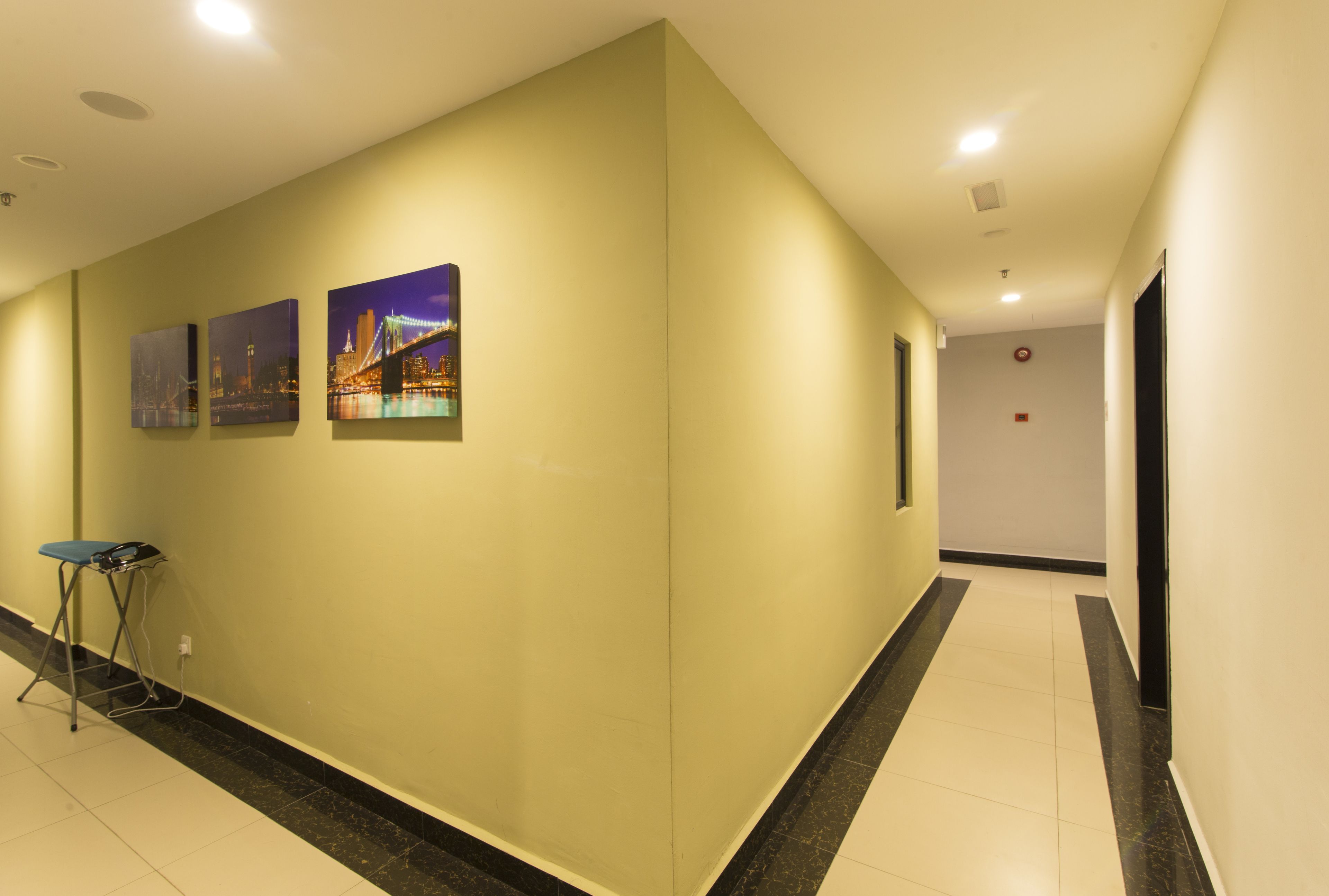 Un pasillo con pared de color amarillo claro, tres cuadros enmarcados, una silla plegable azul y un suelo de baldosas blanco y negro.