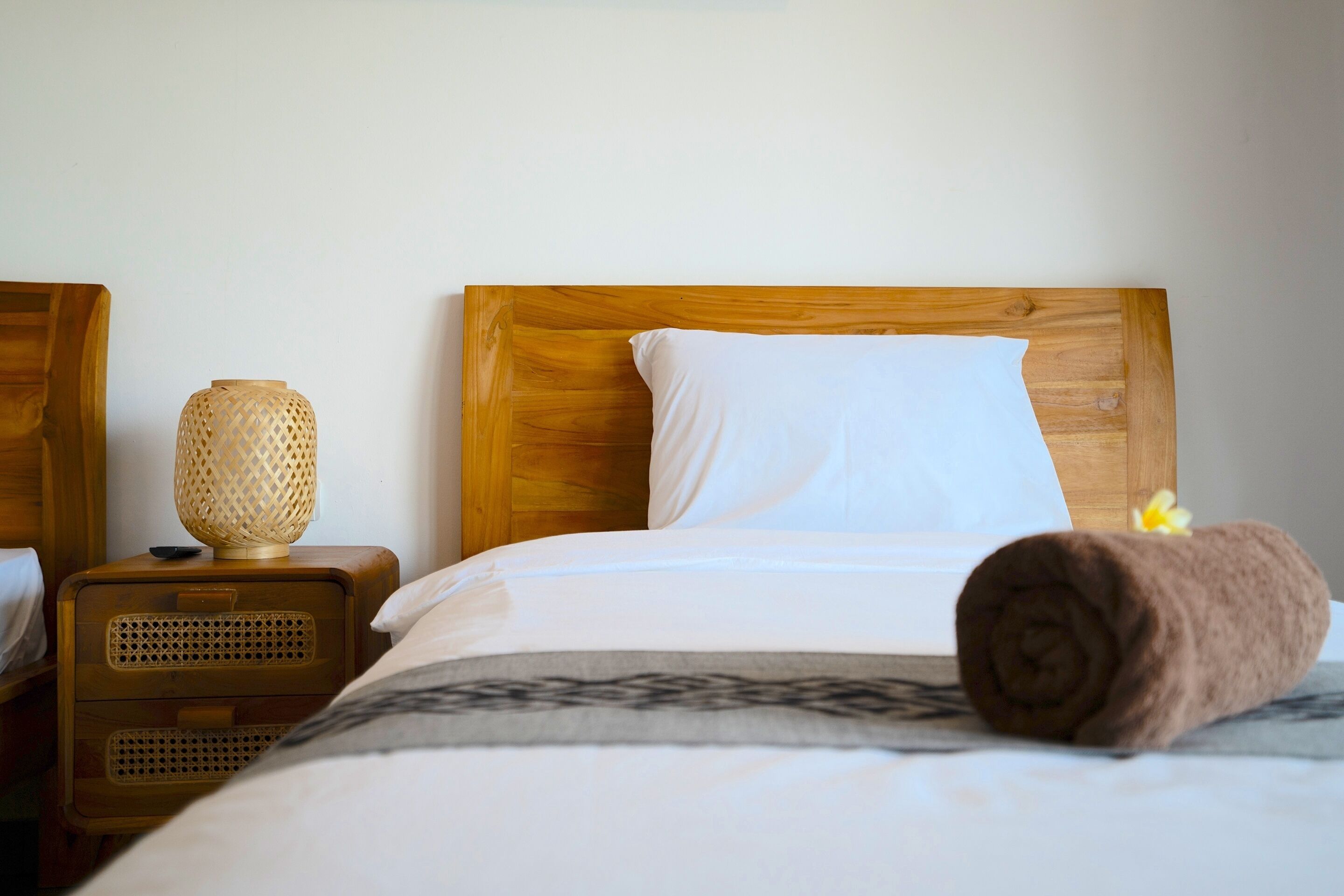 Una cama bien tendida con cabecera de madera, una lámpara de noche y una toalla enrollada sobre la cama.