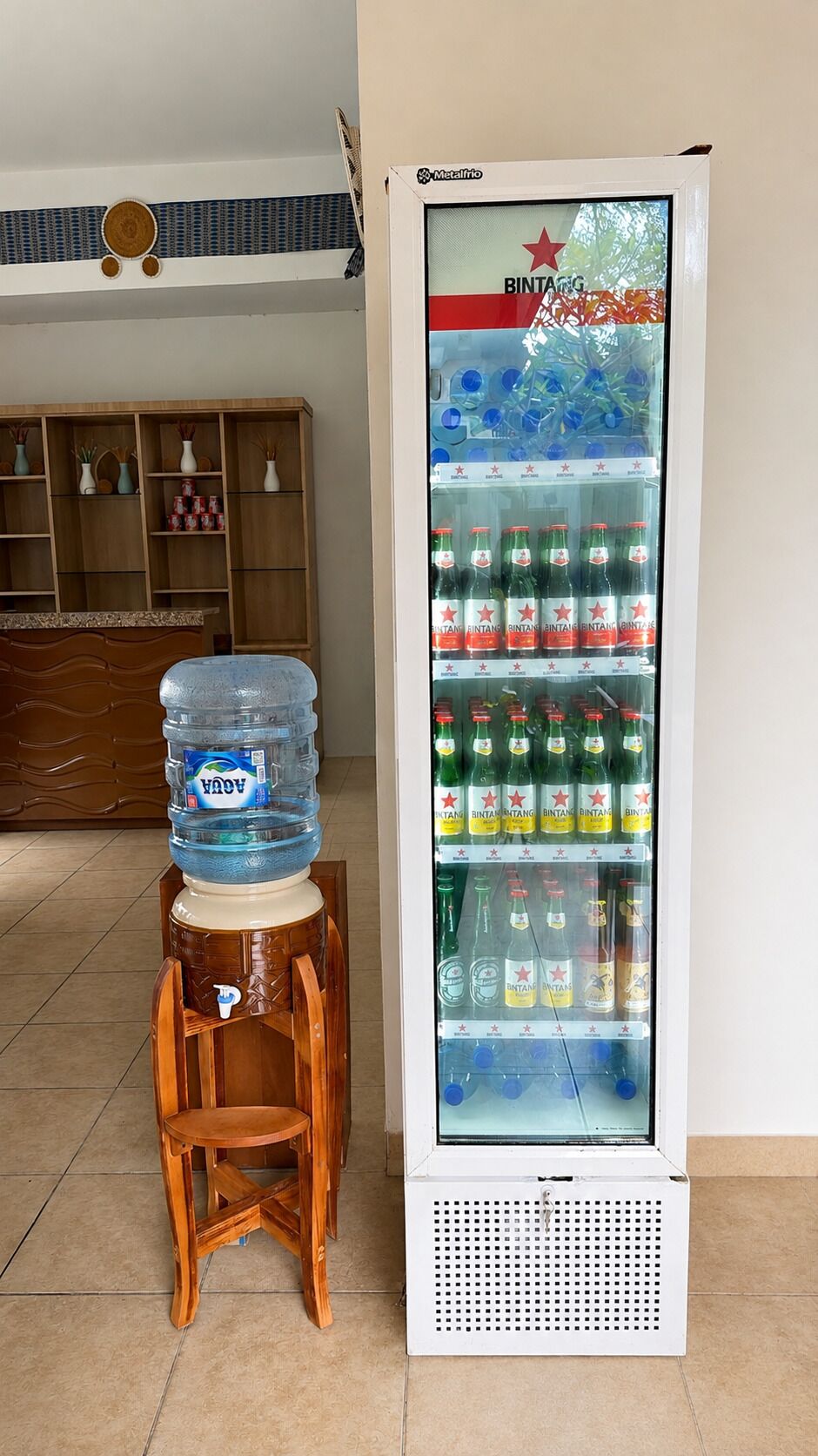 A beverage vending machine with multiple shelves stocked with drinks, a water dispenser on a wooden stool, and a wooden cabinet in the background.