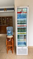 Máquina expendedora de bebidas con varias estanterías llenas de refrescos, un dispensador de agua sobre un taburete de madera y un mueble de madera al fondo.