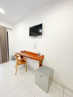 Una habitación pequeña con un escritorio de madera, una silla, un televisor fijado a la pared y un refrigerador pequeño en el suelo.