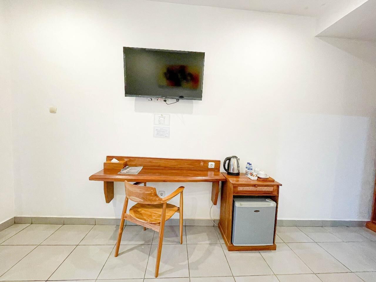 A room with a wooden desk, a chair, a television mounted on the wall, and a small refrigerator.
