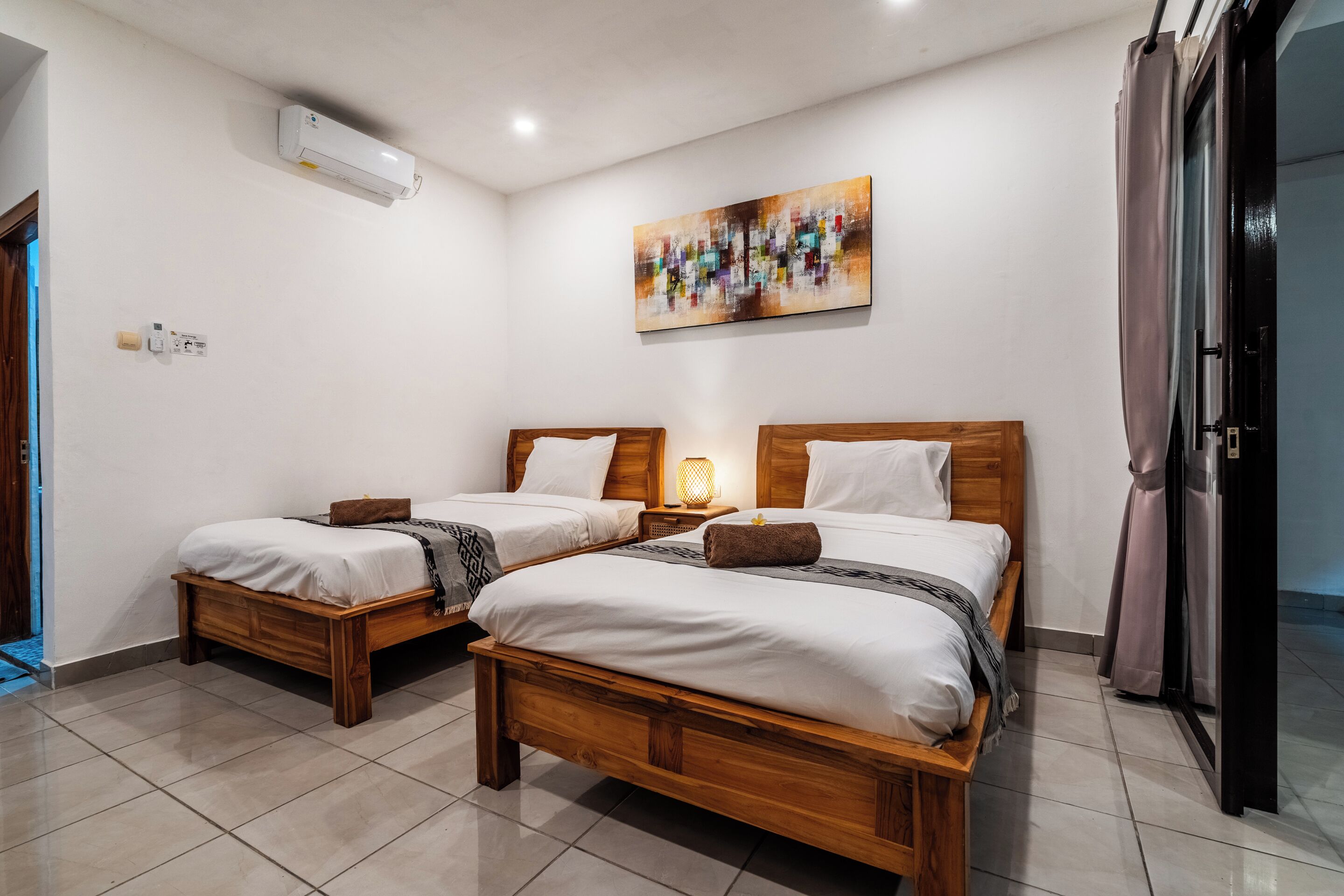 Une chambre d’hôtel avec deux lits en bois, un climatiseur au plafond, un tableau accroché au mur et une porte avec des rideaux.