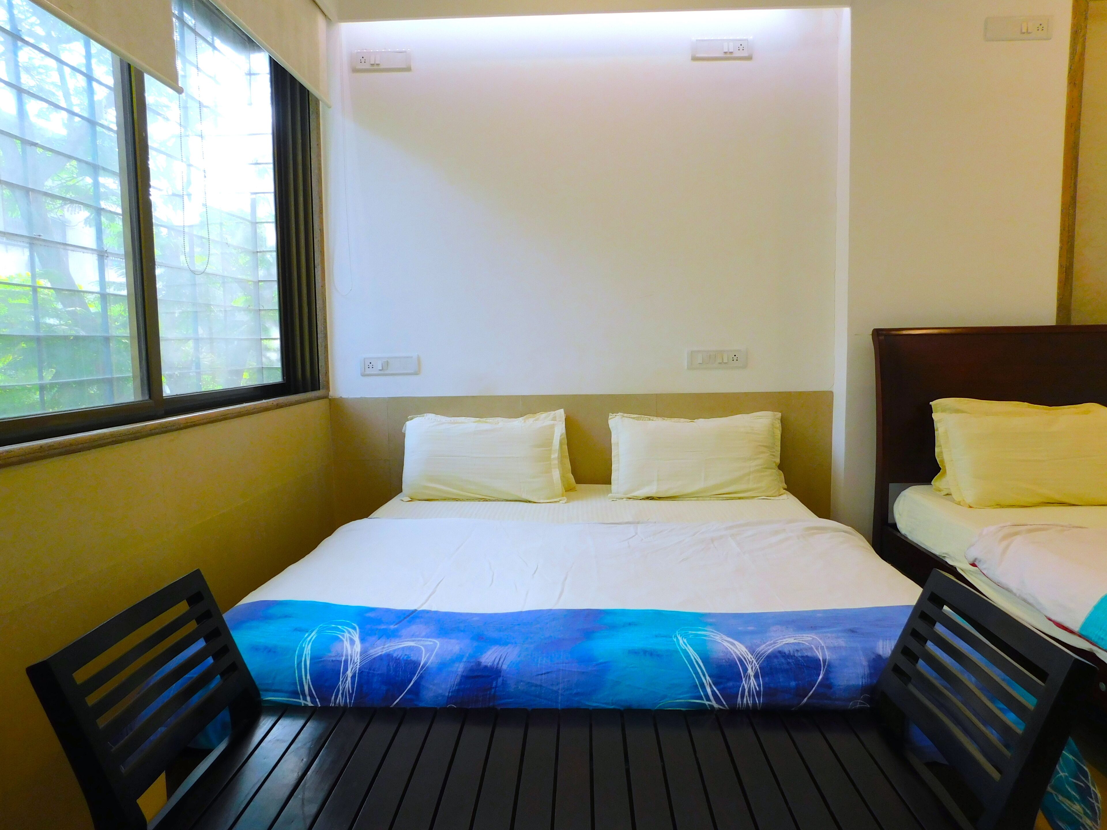 Una habitación pequeña con una cama individual, ventana, enchufe y una silla de madera.