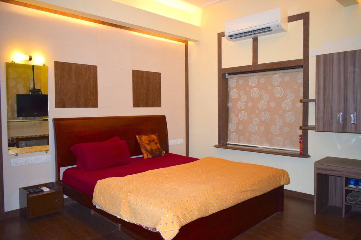 A hotel room with a bed, a bedside table, a mirror, a window with a patterned blind, and a wall-mounted air conditioner.
