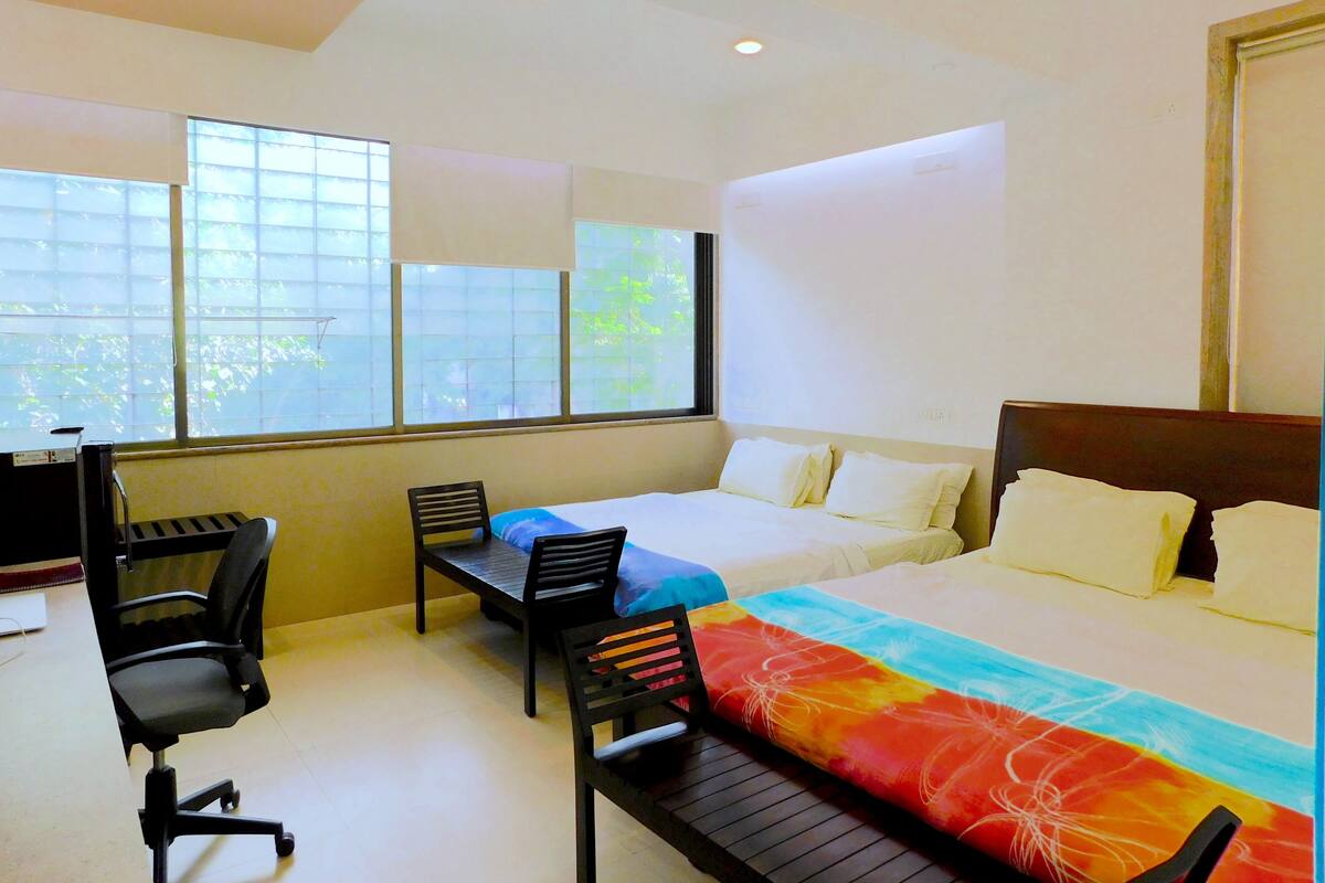 A hotel room with a bed, two chairs, a desk, and large windows.