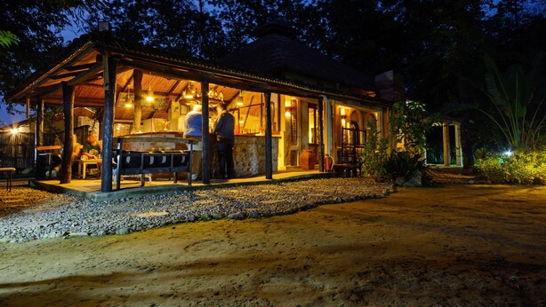 A rustic outdoor restaurant with a thatched roof, wooden tables, and a gravel path leading to it.