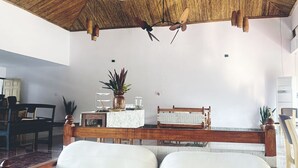 A room with a wooden ceiling, a bed with white bedding, a wooden bench, and a small table with a plant.