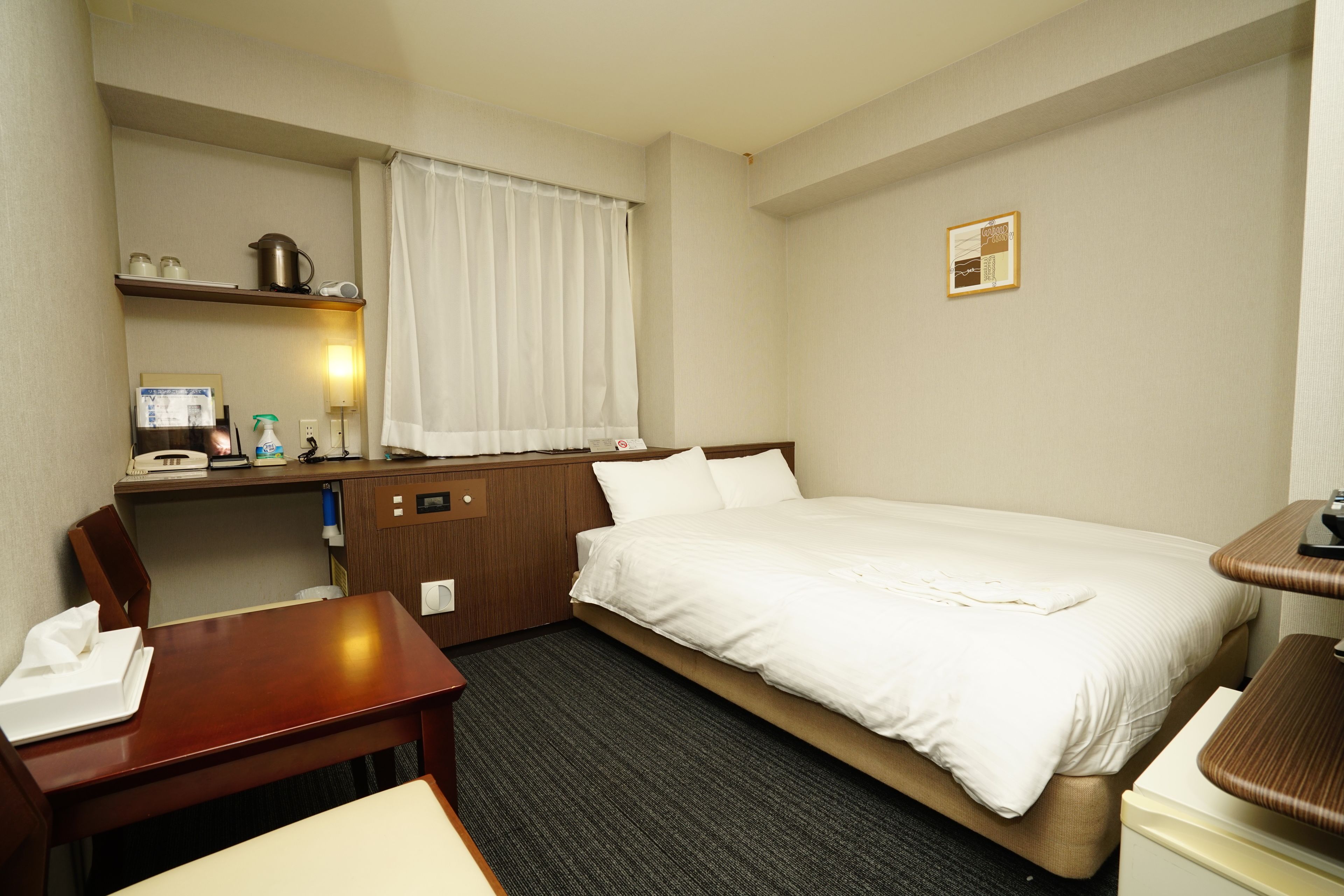 Habitació d'hotel amb un llit, un escriptori, una cadira, una taula petita, una estanteria amb una paperera i cortines a la finestra, i un quadre penjat a la paret.