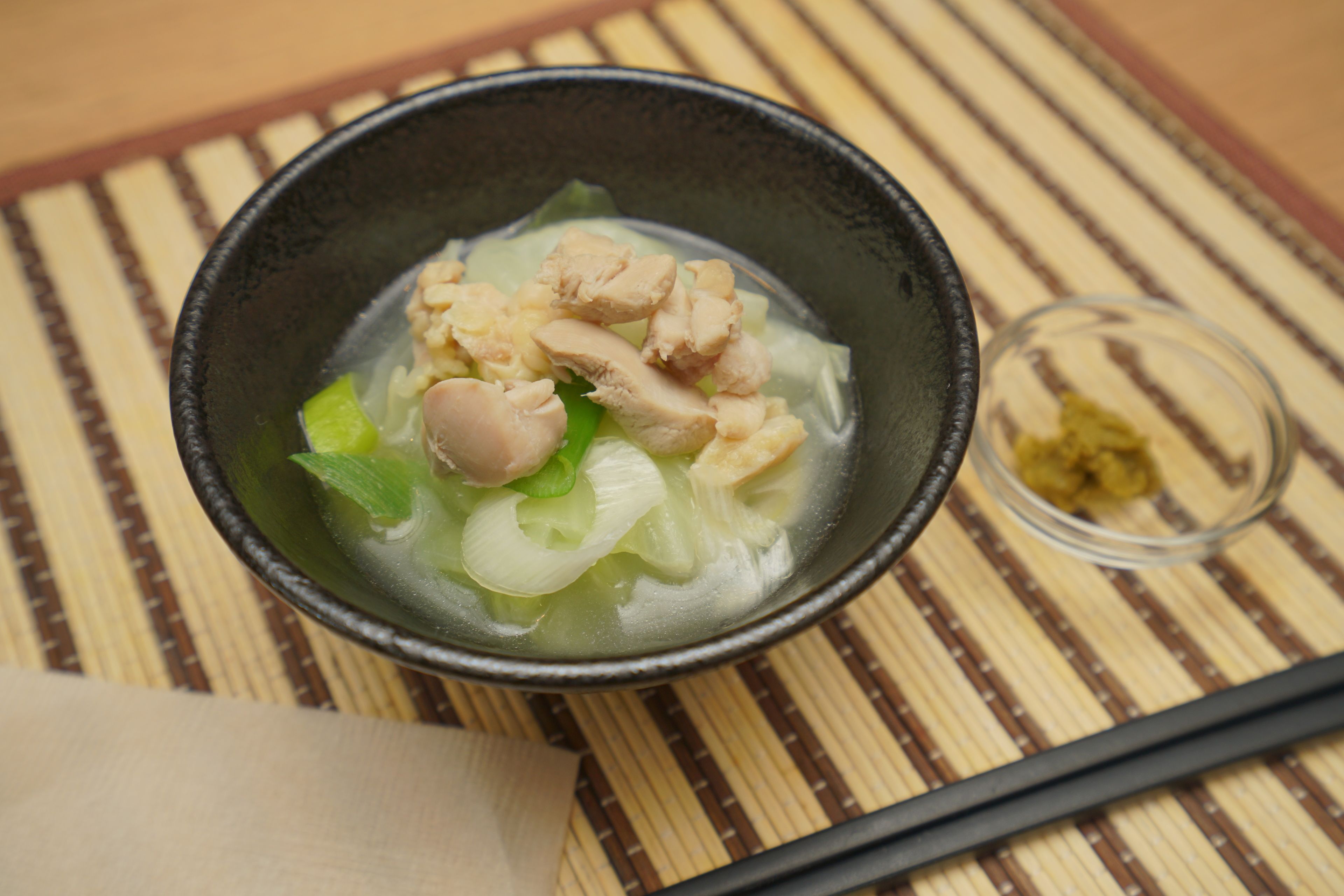 A bowl of soup with vegetables and chicken on a bamboo mat.