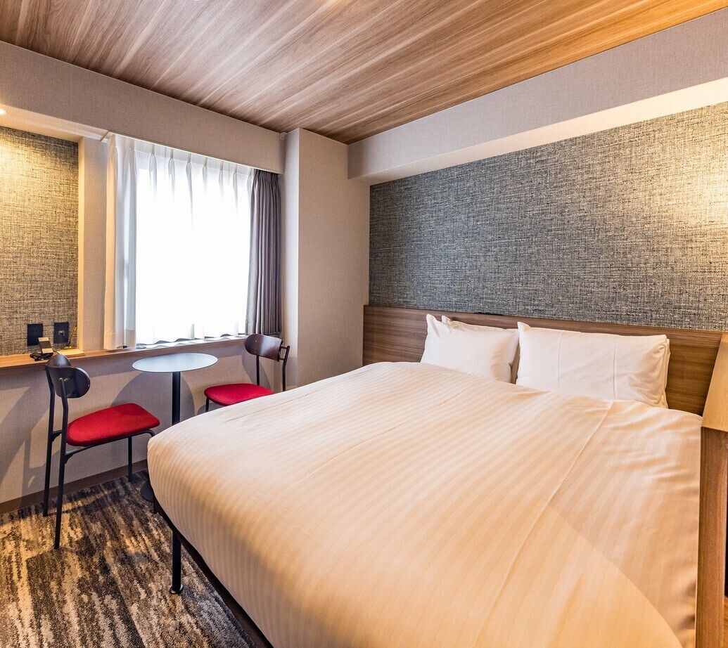 Una habitación de hotel con una cama grande, una mesita pequeña y redonda, dos sillas rojas, una ventana con cortinas y una pared texturizada detrás de la cama.