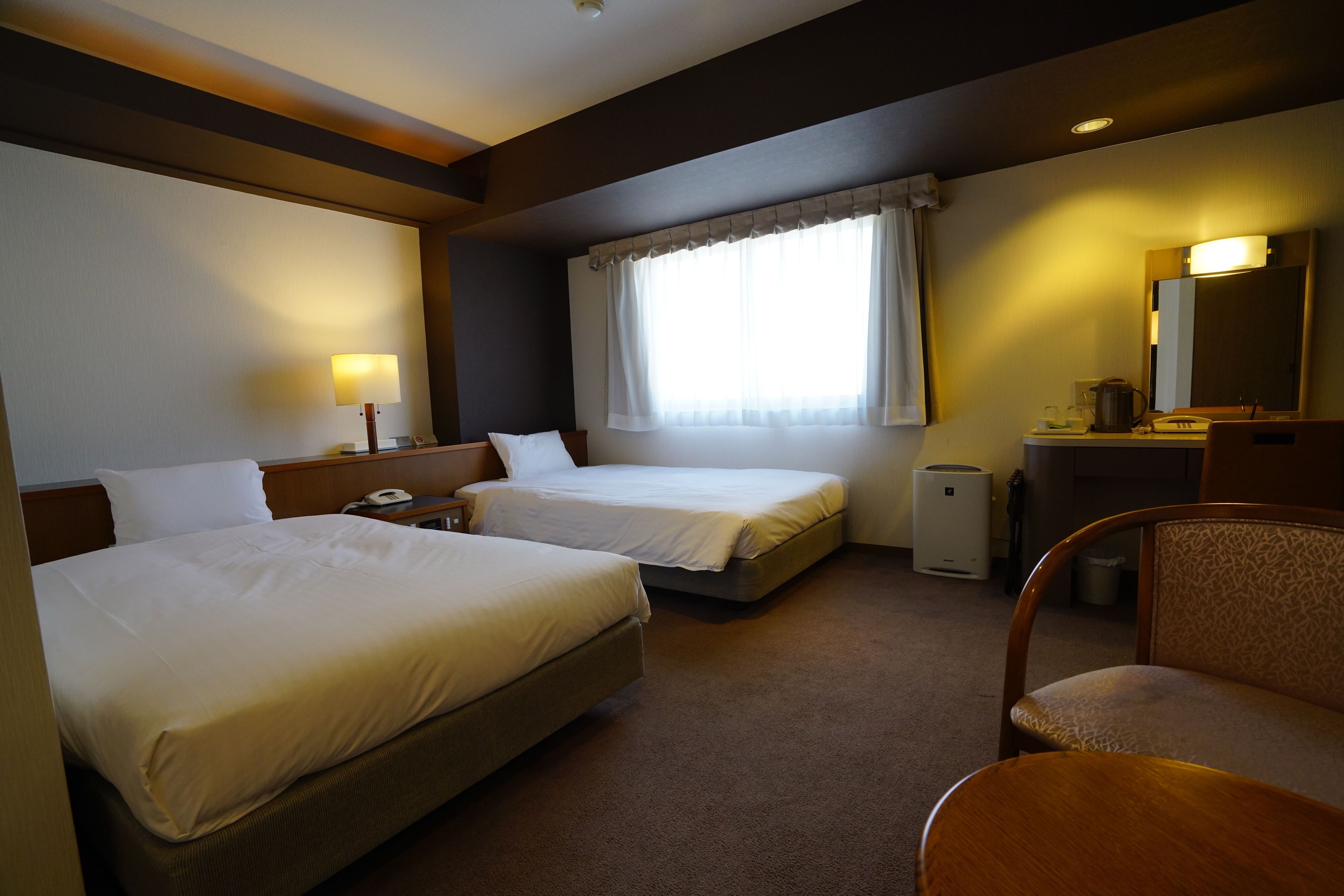 A hotel room with two single beds, a desk with a lamp, a chair, a small table, a mirror, and a window with curtains.