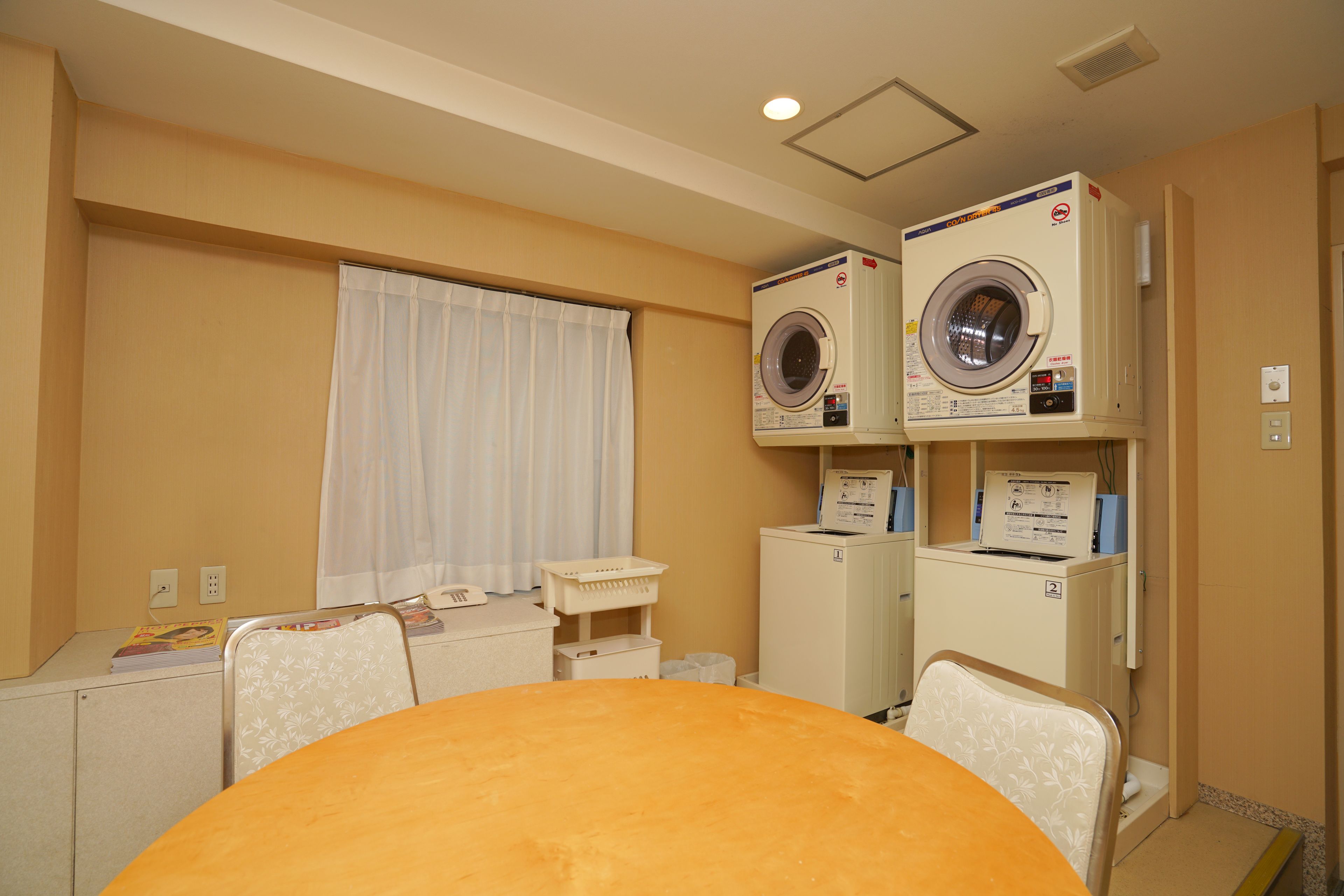 A hotel room with a bed, a table, a chair, a window with curtains, and two stacked washing machines.