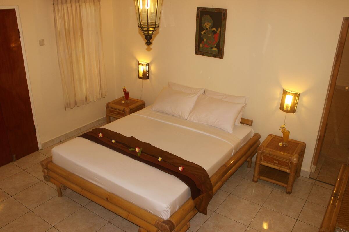 A neatly made bed with a wooden frame, two bedside tables with lamps, a window with curtains, and a wall-mounted picture.