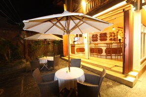 An outdoor patio with wicker chairs and a table, an umbrella, and a building entrance with a bar visible in the background. - Chonos Hotel Lovina (Buleleng)