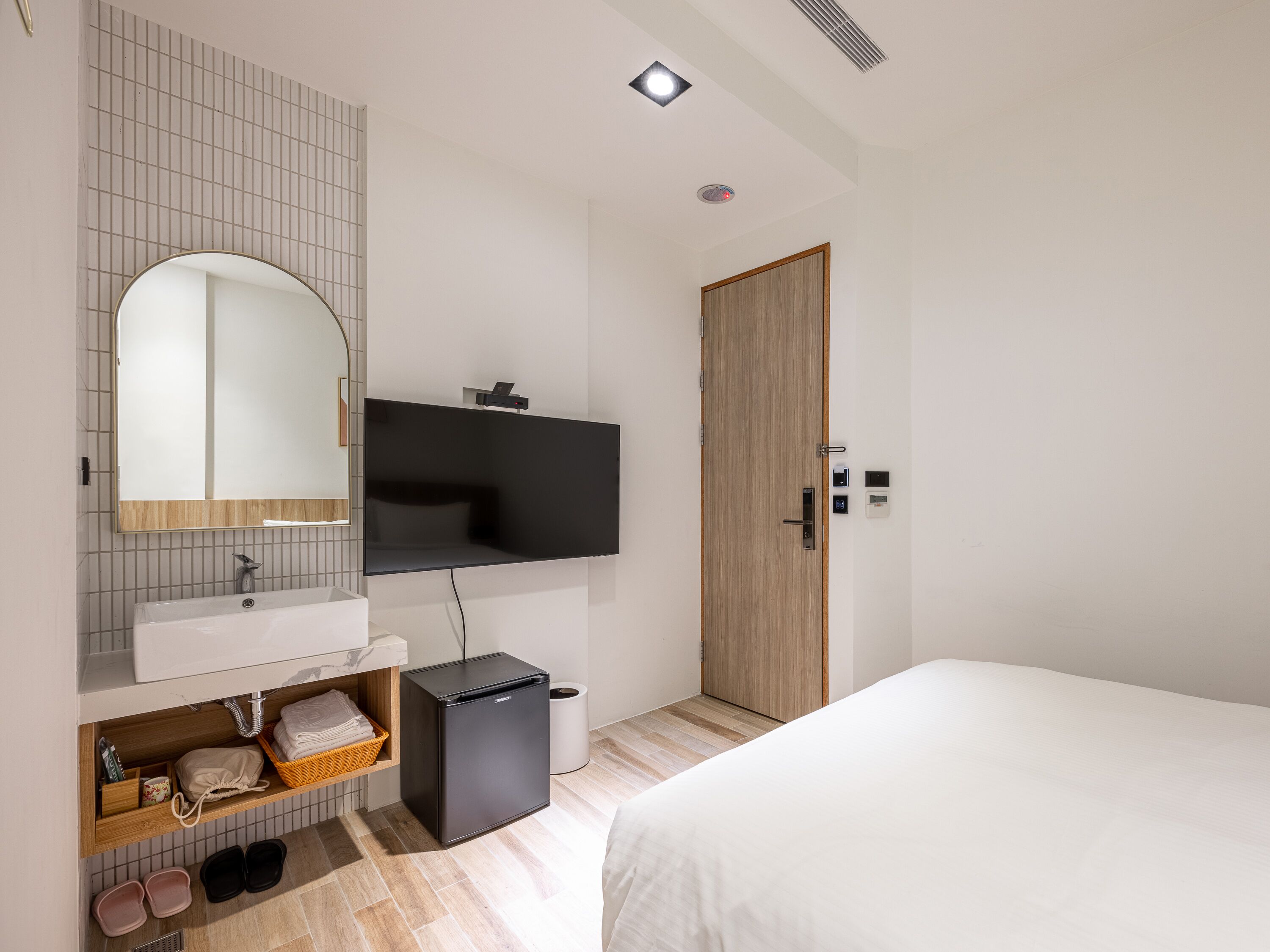 Una habitación de hotel moderna con una cama grande, televisión integrada, armario de madera, baño con lavamanos y espejo, y un refrigerador pequeño.