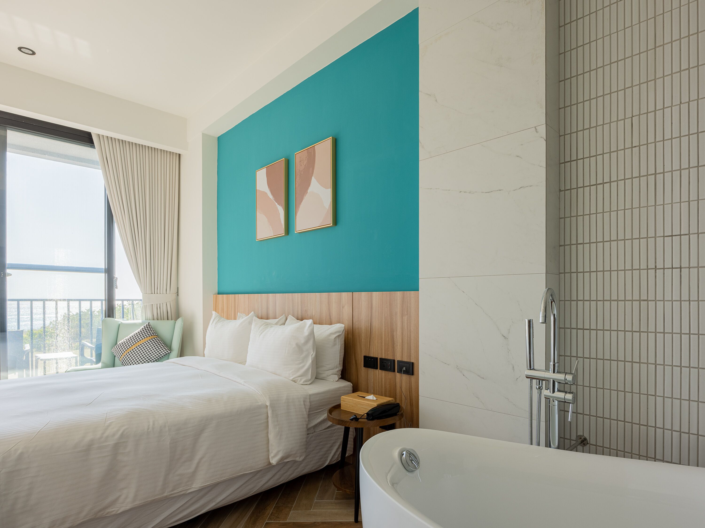 A modern hotel room with a large bed, a wooden headboard, a bathtub, and a view of the sea.