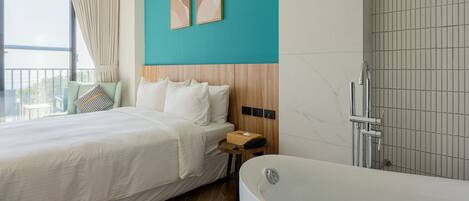 A modern hotel room with a large bed, a wooden headboard, a bathtub, and a view of the sea.