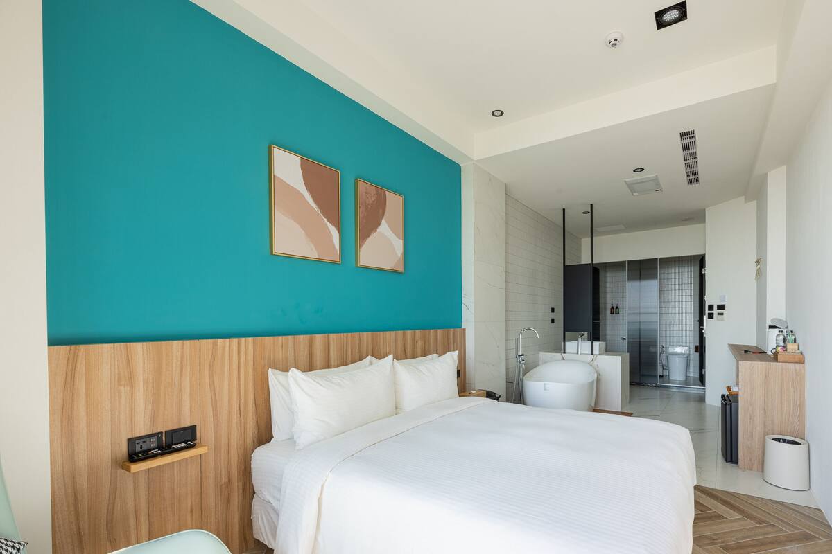 Ett modernt hotellrum med en stor säng, en sänggavel i trä, en vägg i en turkos nyans och ett badrum med badkar och toalett.