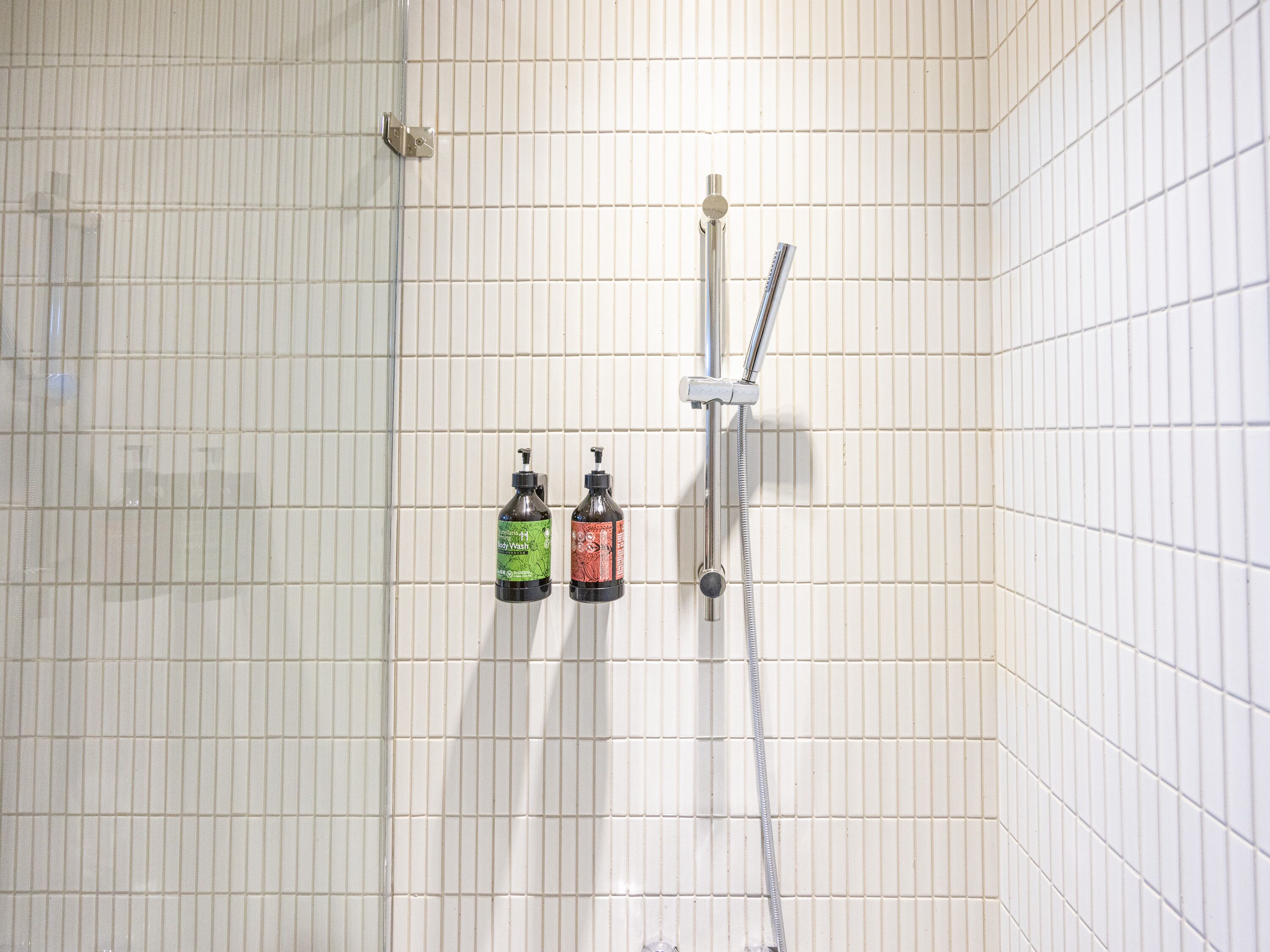 Un área de ducha con azulejos, un cabezal de ducha de mano y dos dispensadores de jabón.