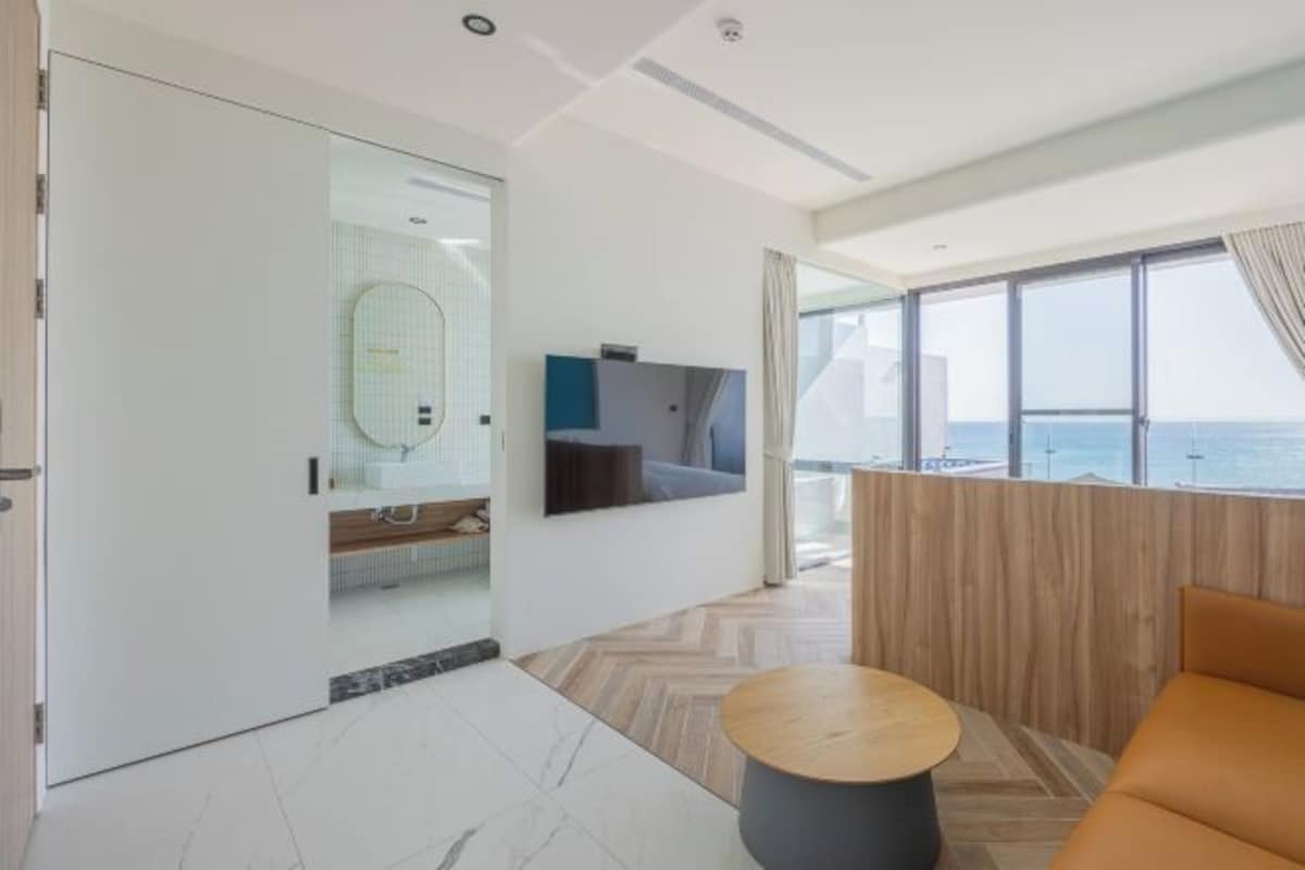 A modern hotel room with a large window offering a view of the sea, a wooden wall, and a seating area with a sofa and a round table.