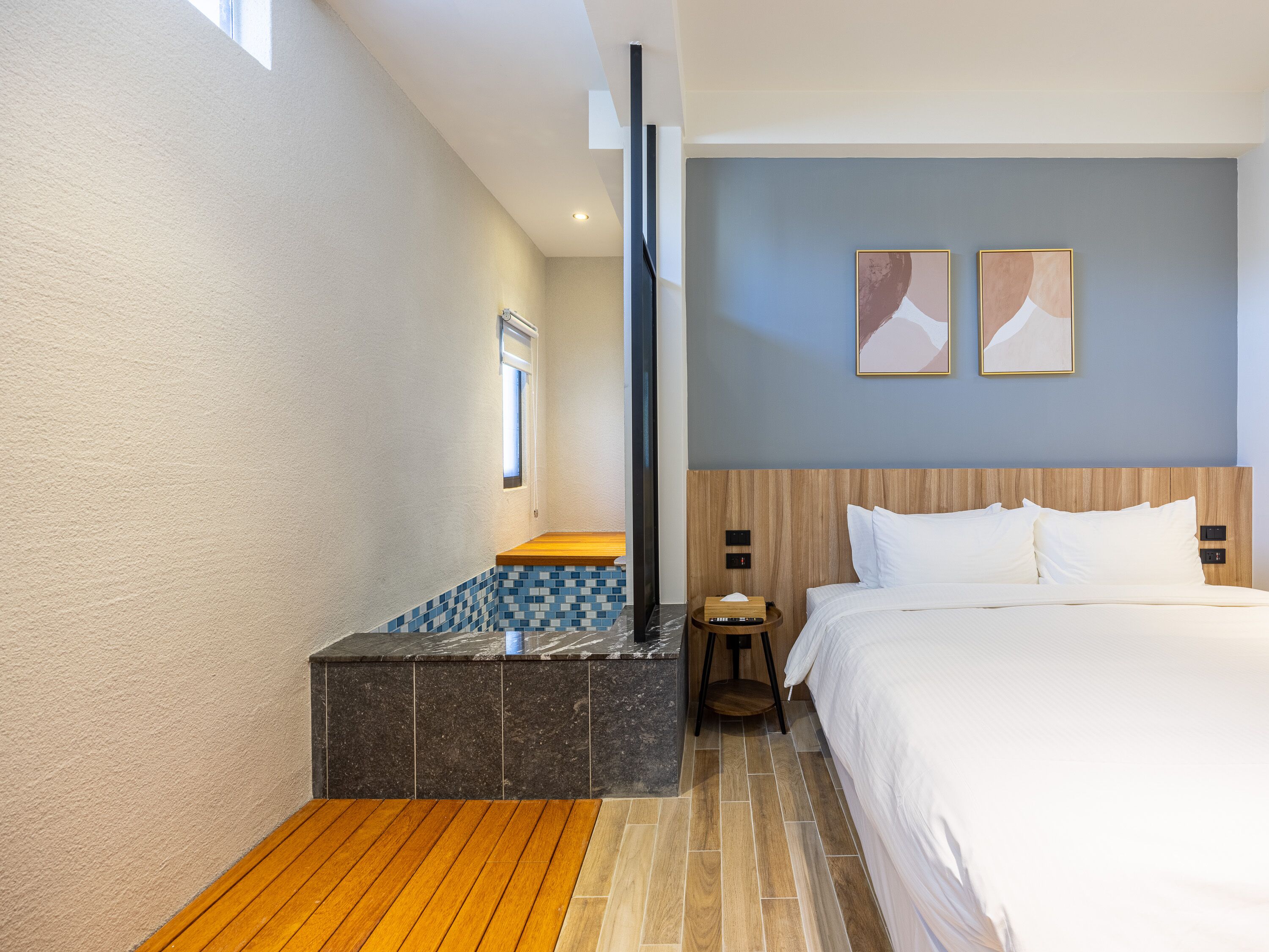 A modern hotel room with a wooden bench, a bed with white linens, a nightstand, and two abstract paintings on the wall.