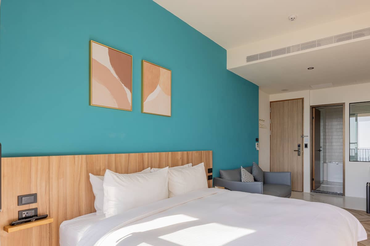Ett modernt hotellrum med en stor säng, en sänggavel i trä och väggar i en nyans av turkos. Ovanför sängen hänger två inramade, abstrakta målningar.
