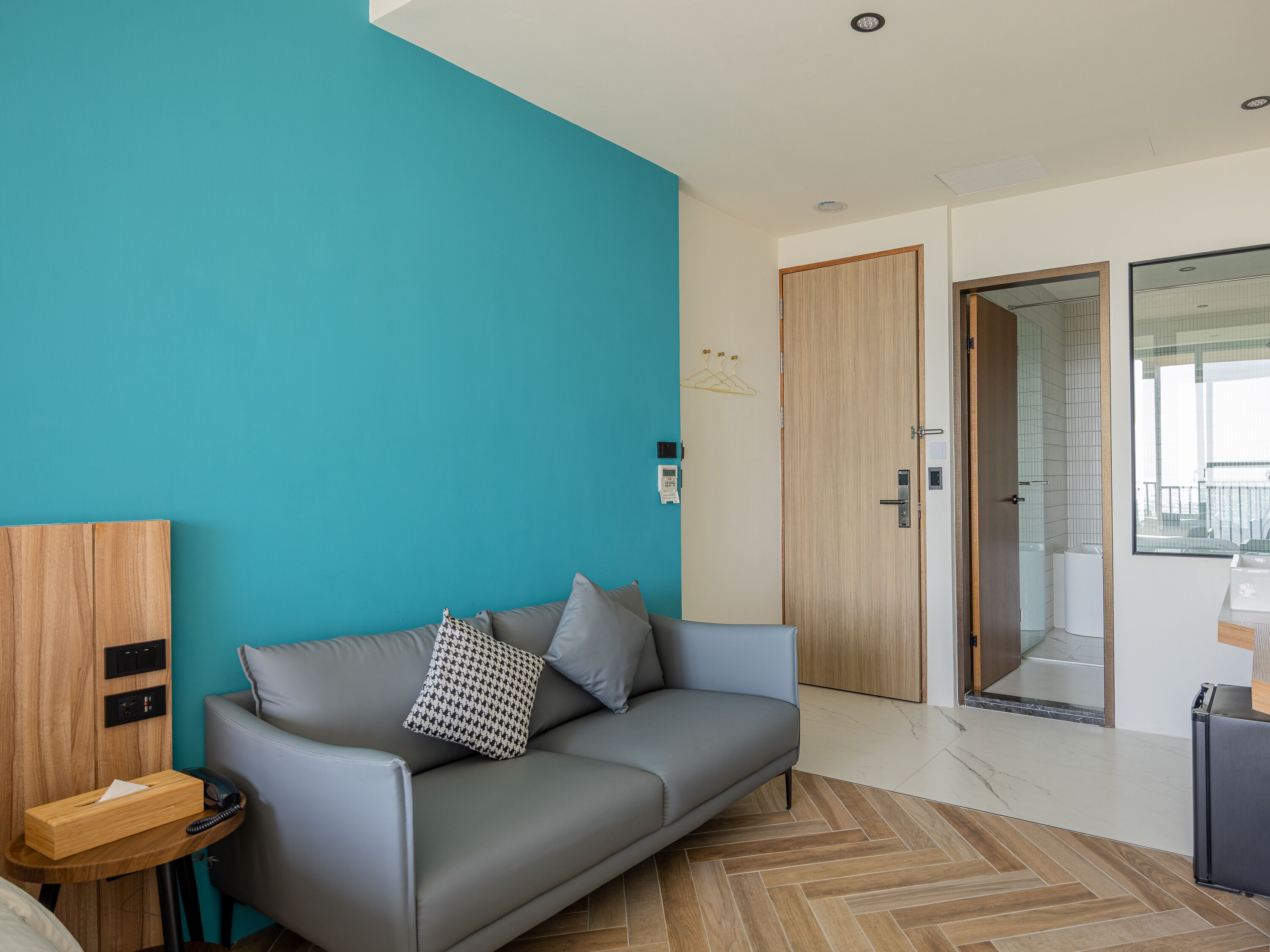 A modern hotel room with a grey sofa, a wooden side table, a wooden door, and a bathroom visible through a glass door.