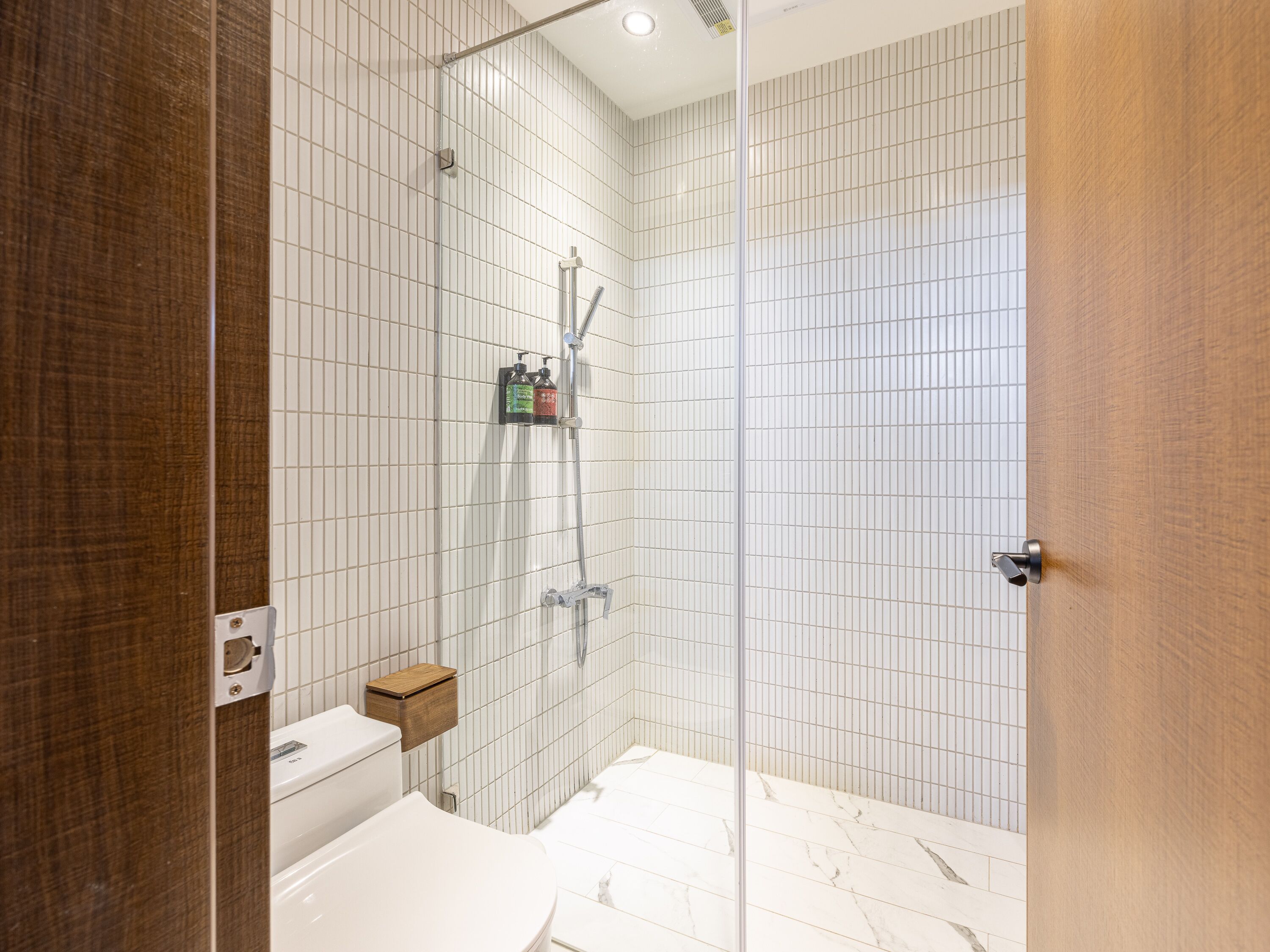 A modern bathroom with a glass shower enclosure, white tiled walls, and a wooden door.