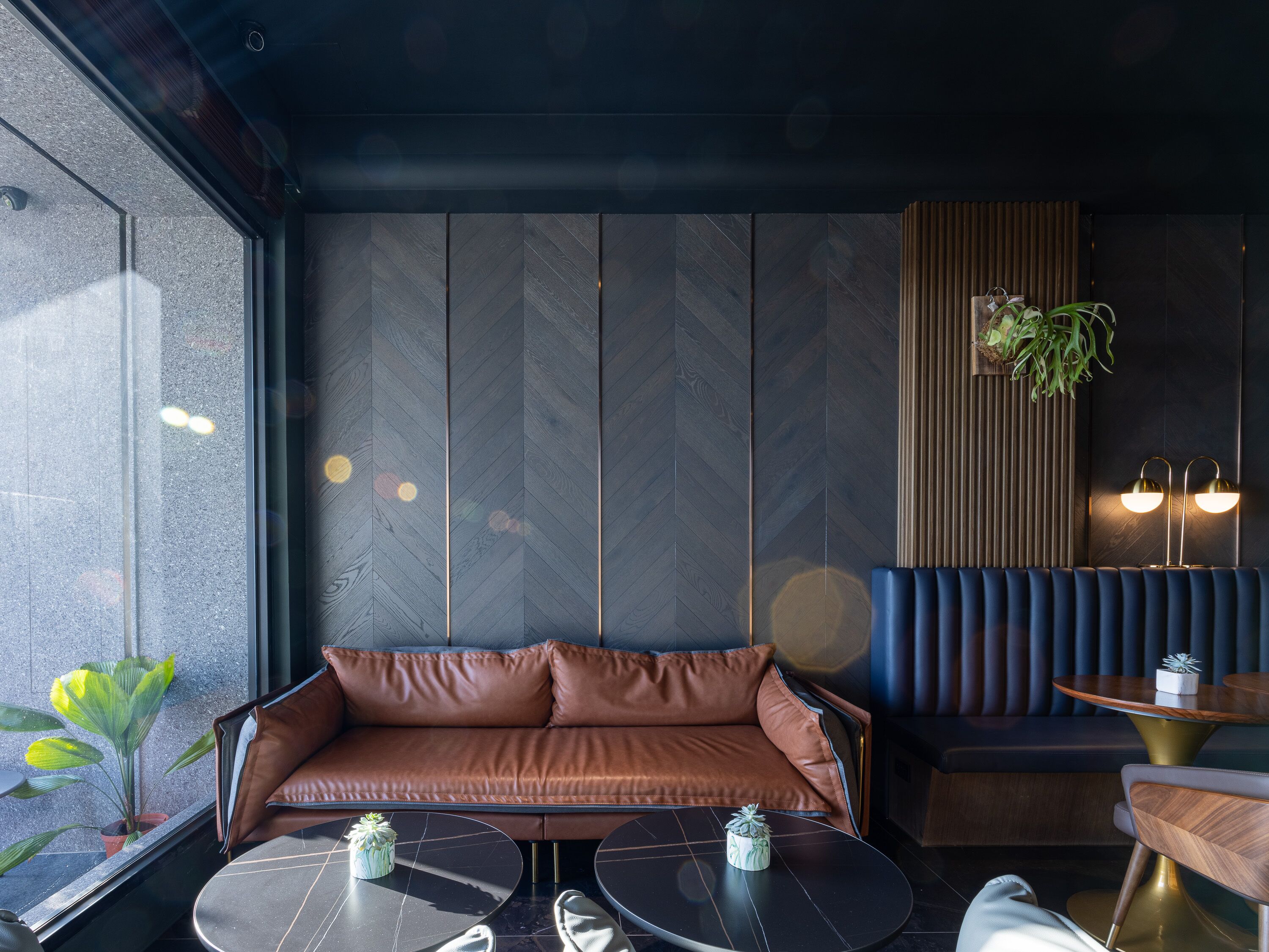 A modern interior with a brown leather sofa, two round tables with small plants, a wooden bench, and a large window with a view.
