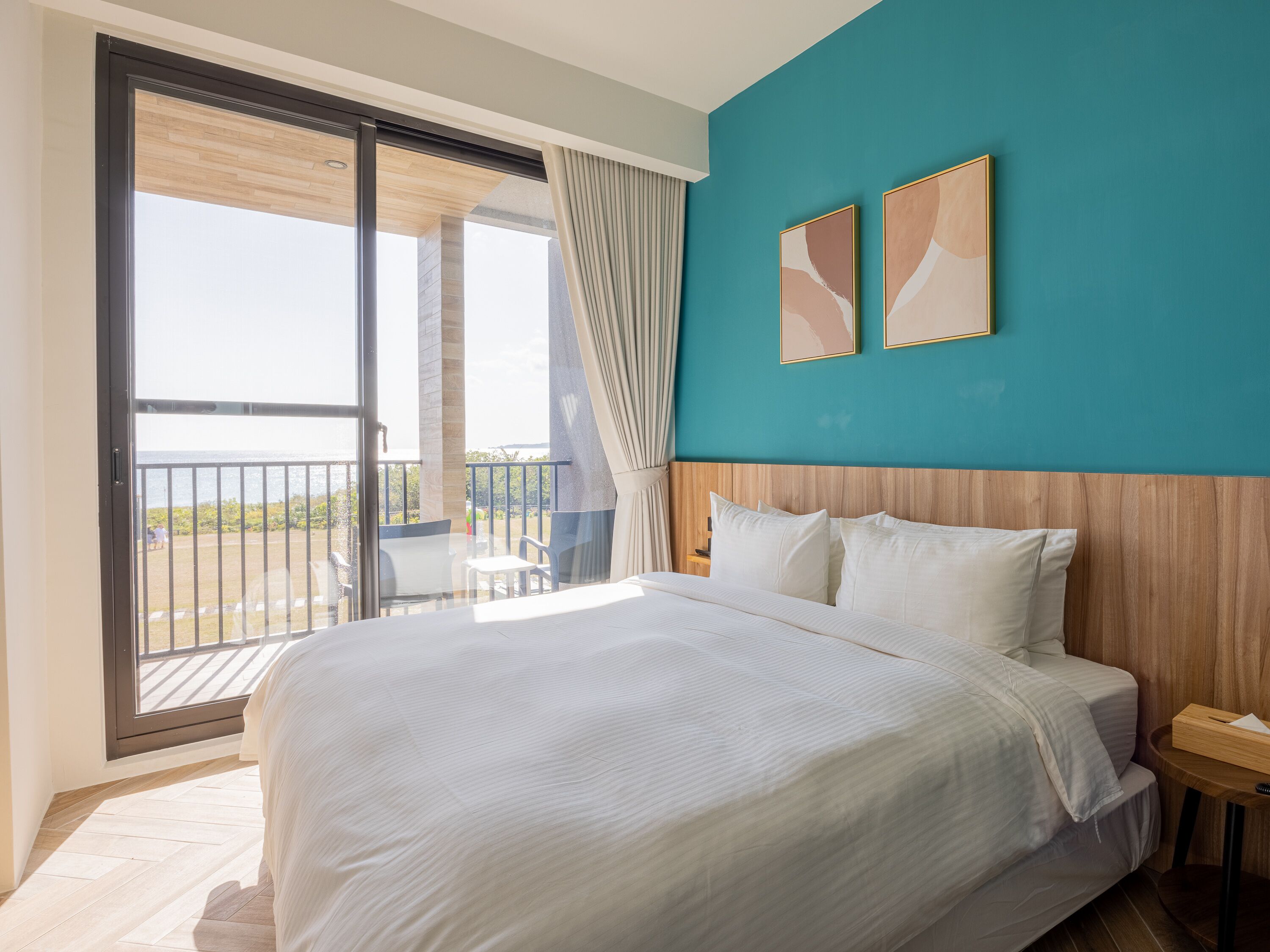 A hotel room with a large bed, a wooden headboard, a view of the sea through a sliding glass door, and two framed artworks on the teal-colored wall.