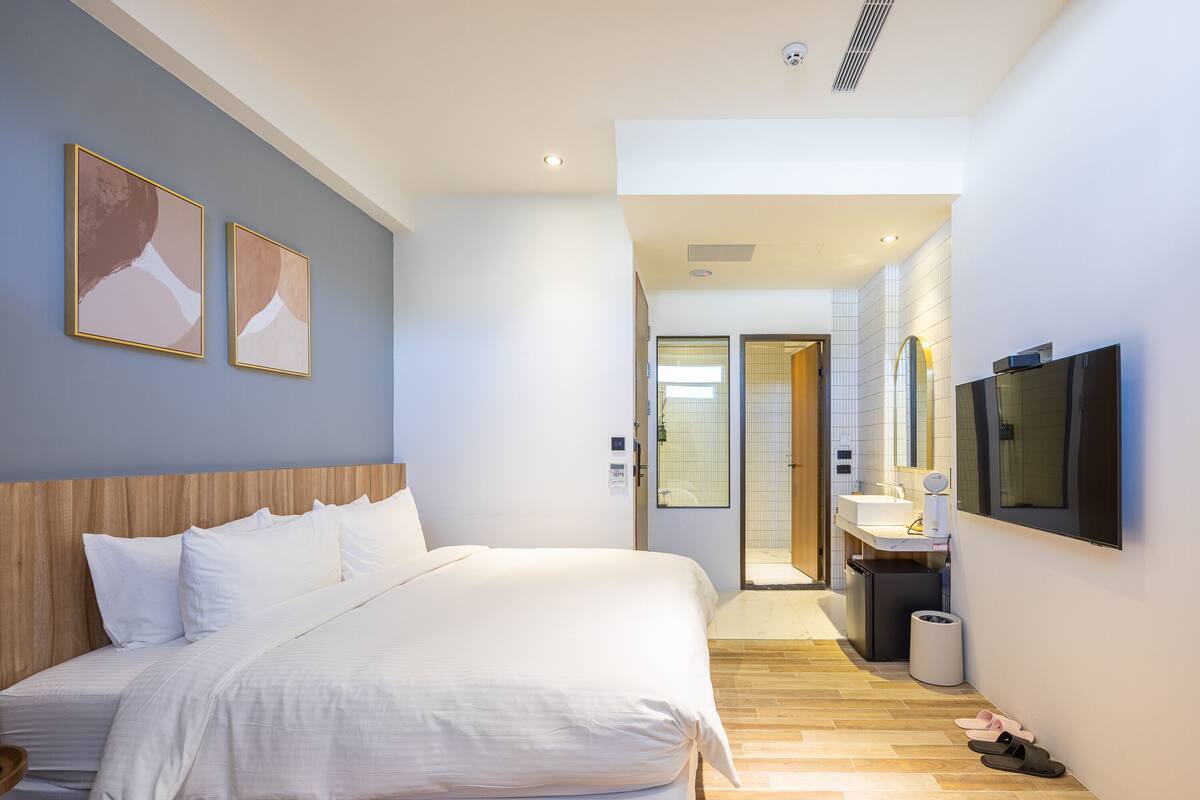 Ett modernt hotellrum med en stor säng, en platt-TV som är monterad på väggen och en duschhörna som syns genom en öppen dörr.