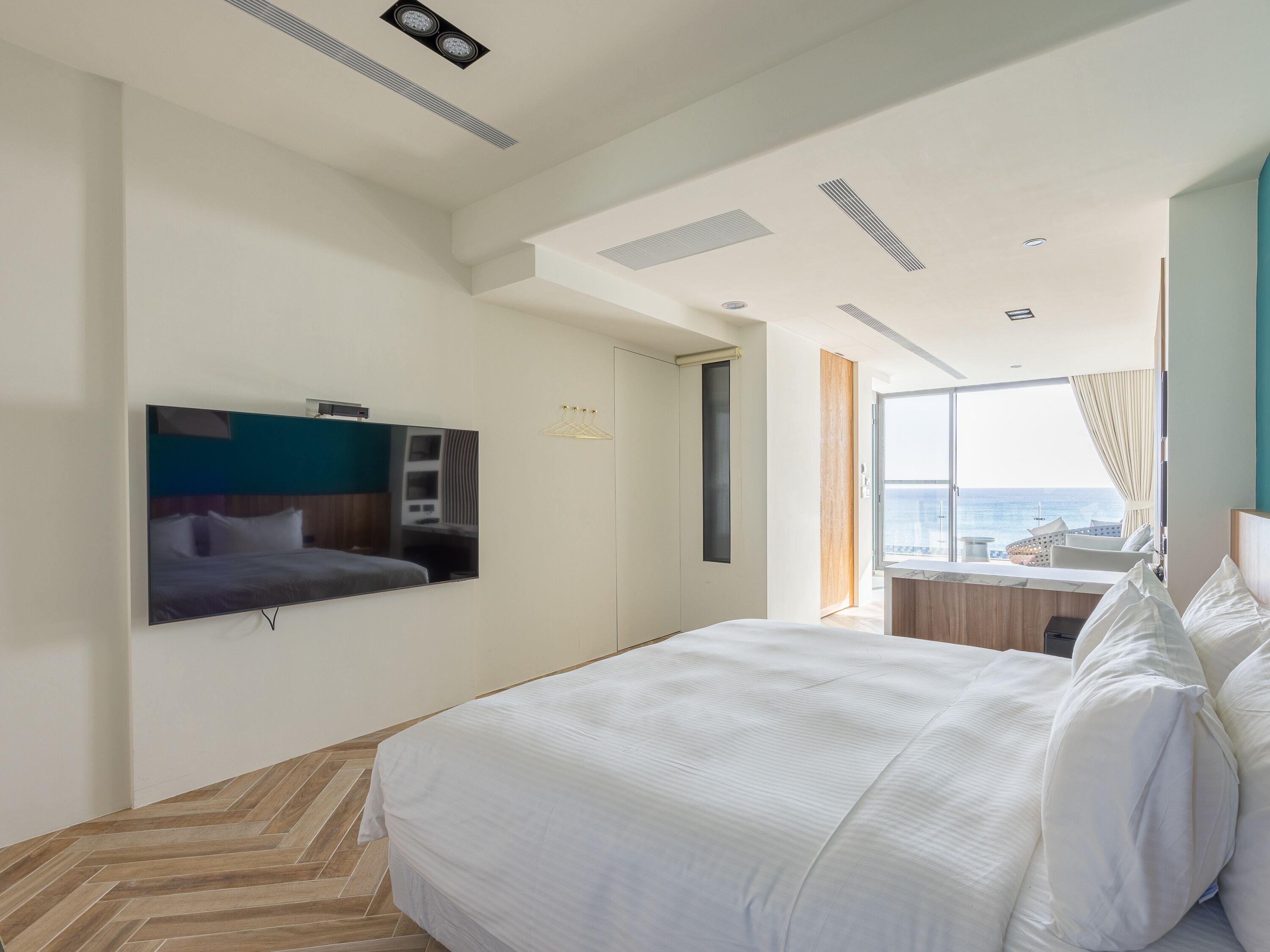 A modern hotel room with a large bed, a flat-screen TV mounted on the wall, a window with a view of the sea, and a balcony with seating.