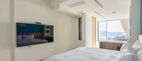 A modern hotel room with a large bed, a flat-screen TV mounted on the wall, a window with a view of the sea, and a balcony with seating.