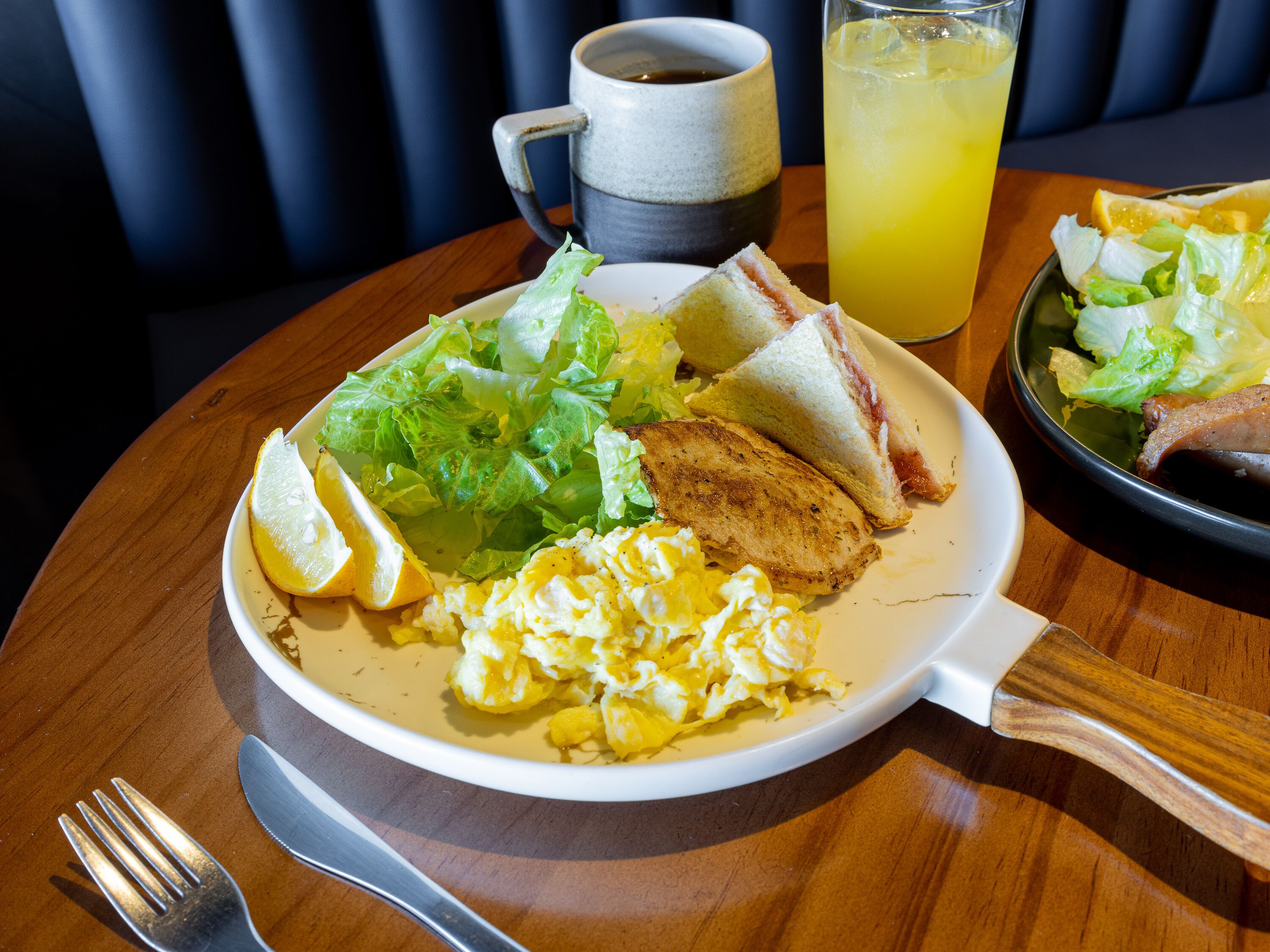Une assiette de petit-déjeuner avec des œufs, des toasts et une salade, une tasse et un verre de jus d’orange, le tout posé sur une table en bois.