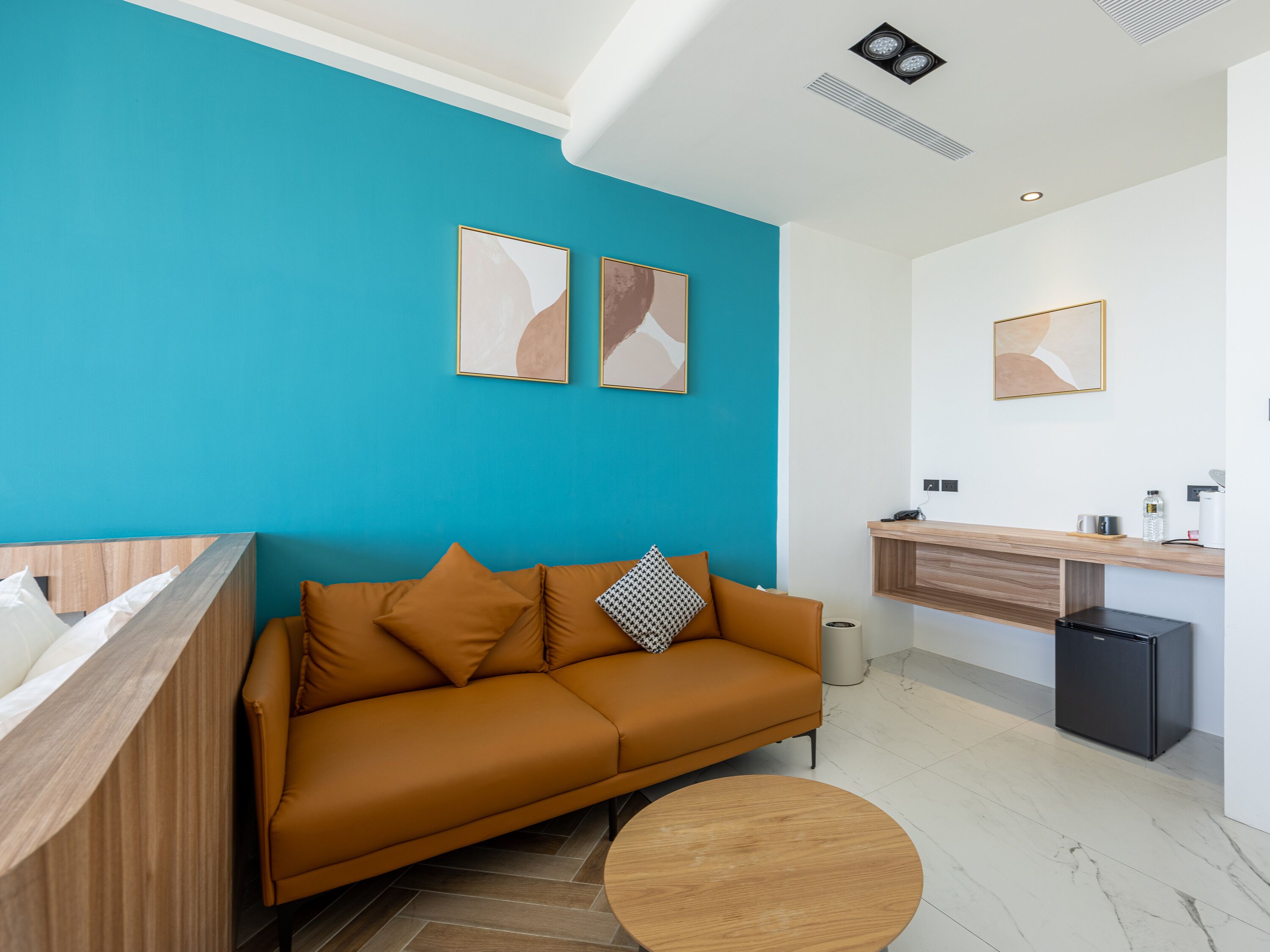 A modern living room with a teal accent wall, a wooden sofa, a round coffee table, and a kitchenette area with a refrigerator and sink.